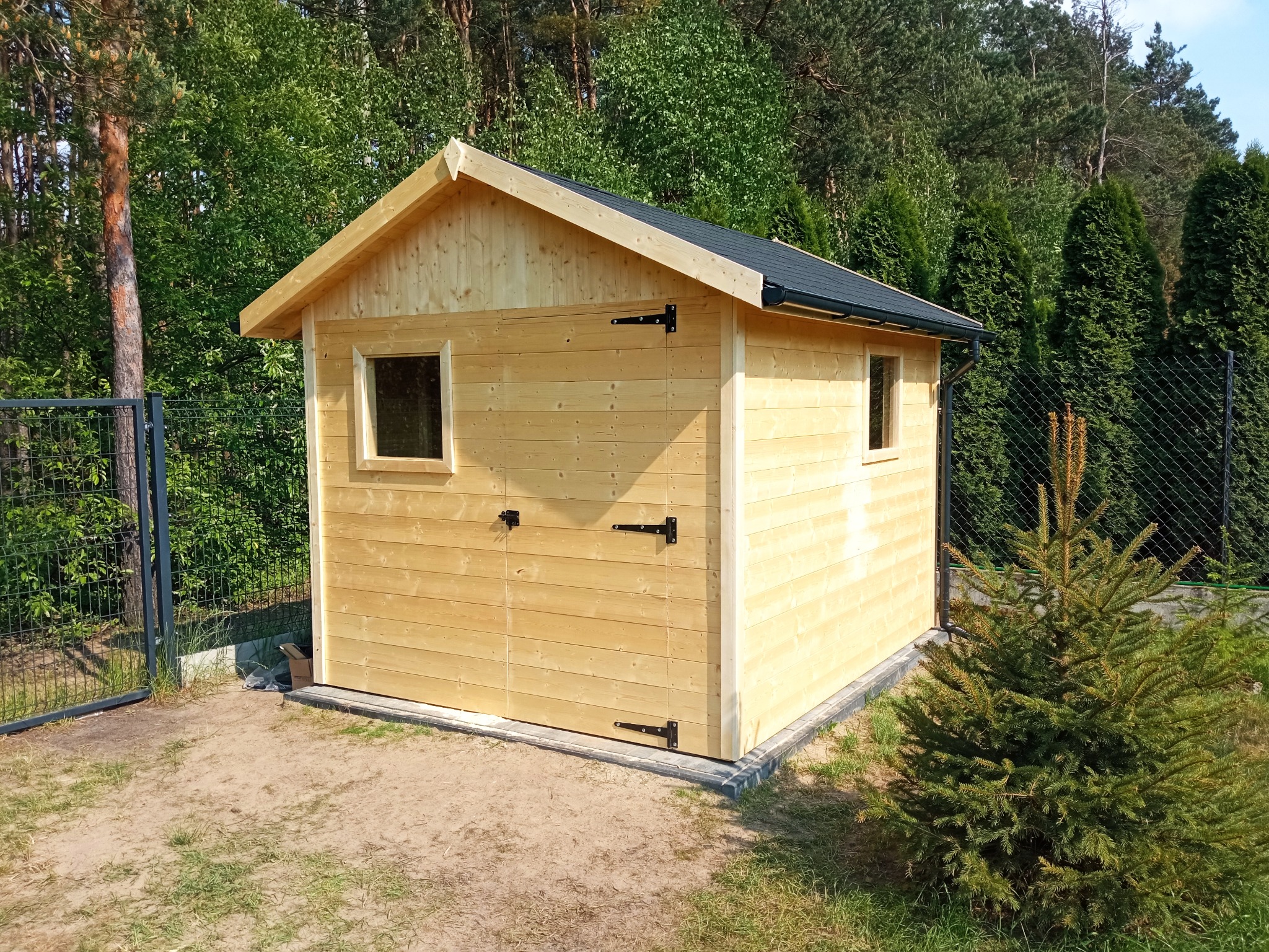 Niewielki, surowy, drewniany domek narzędziowy z czarnym, spadzistym dachem i dwoma oknami, otoczony zielenią i ogrodzeniem z siatki.