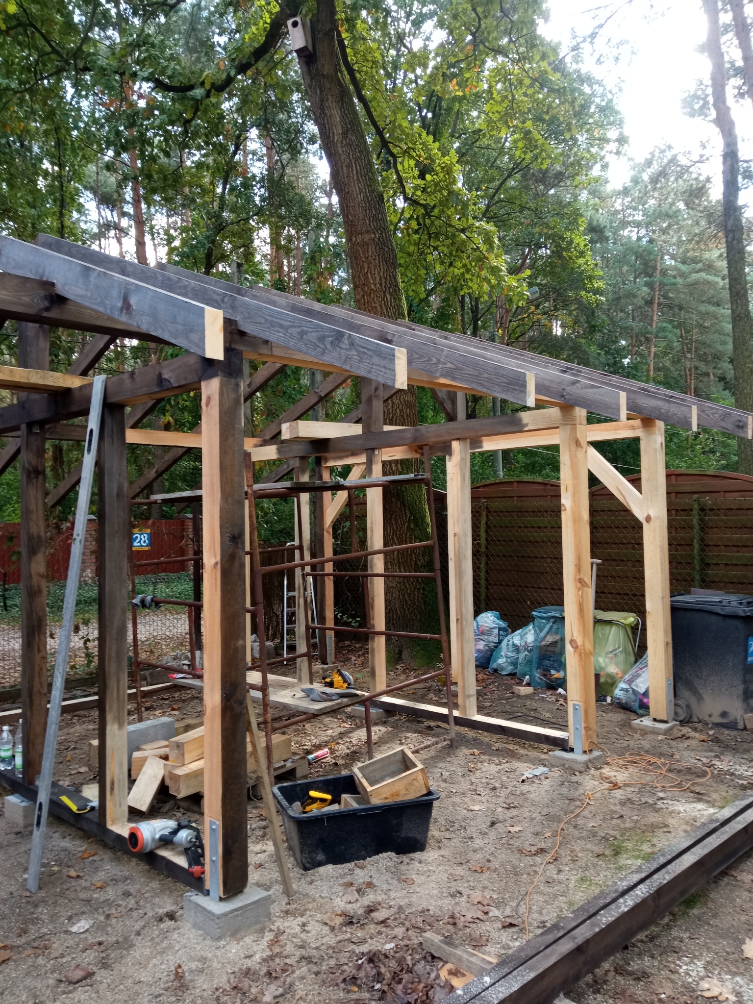 Konstrukcja drewniana w trakcie budowy, widoczne narzędzia, poziomica oparta o słup, w tle drzewa i ogrodzenie.