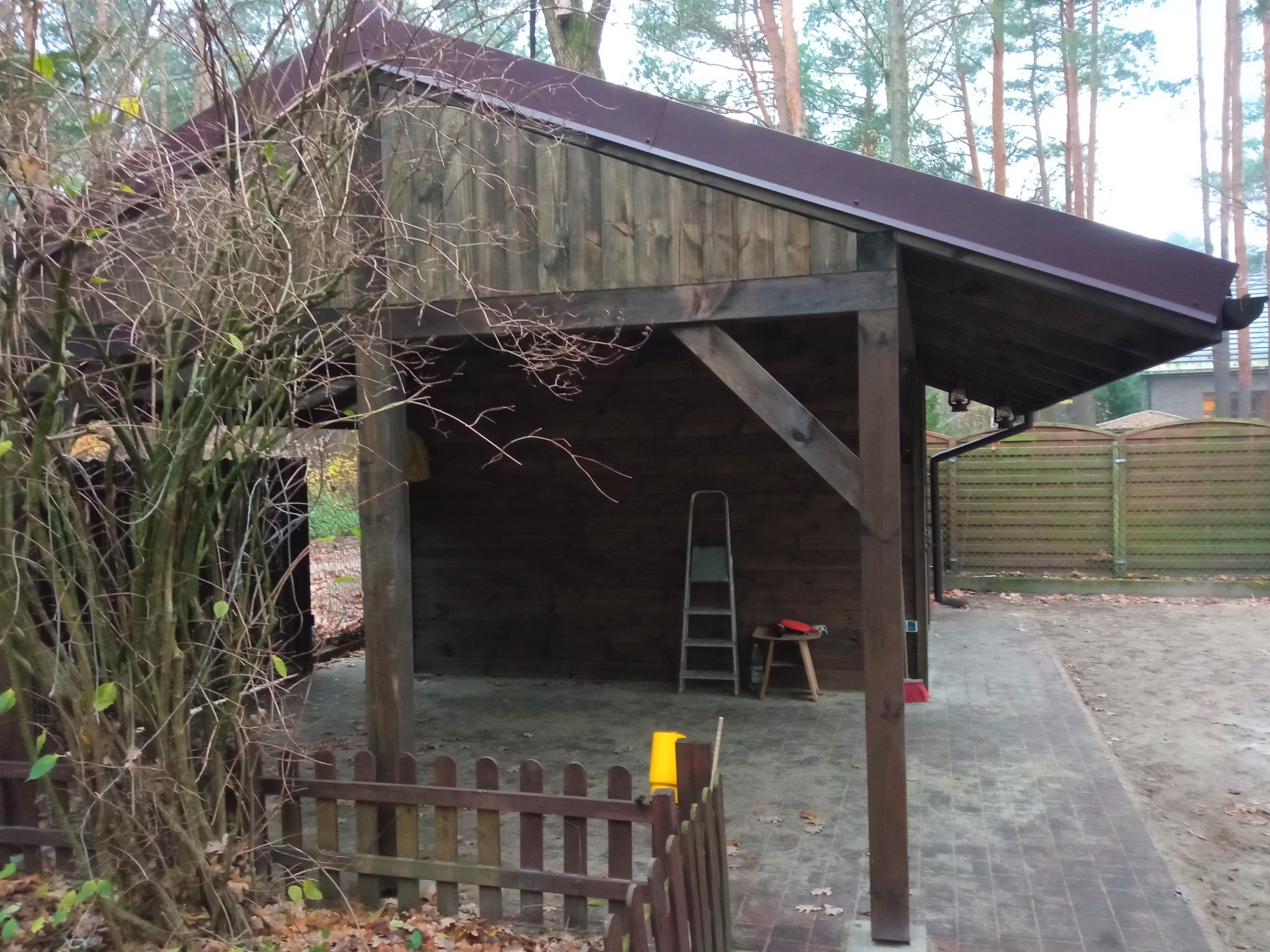 Drewniana konstrukcja z dachem z blachy, prawdopodobnie zadaszenie lub wiata, widziana z perspektywy niskiego ogrodzenia w jesiennej scenerii.
