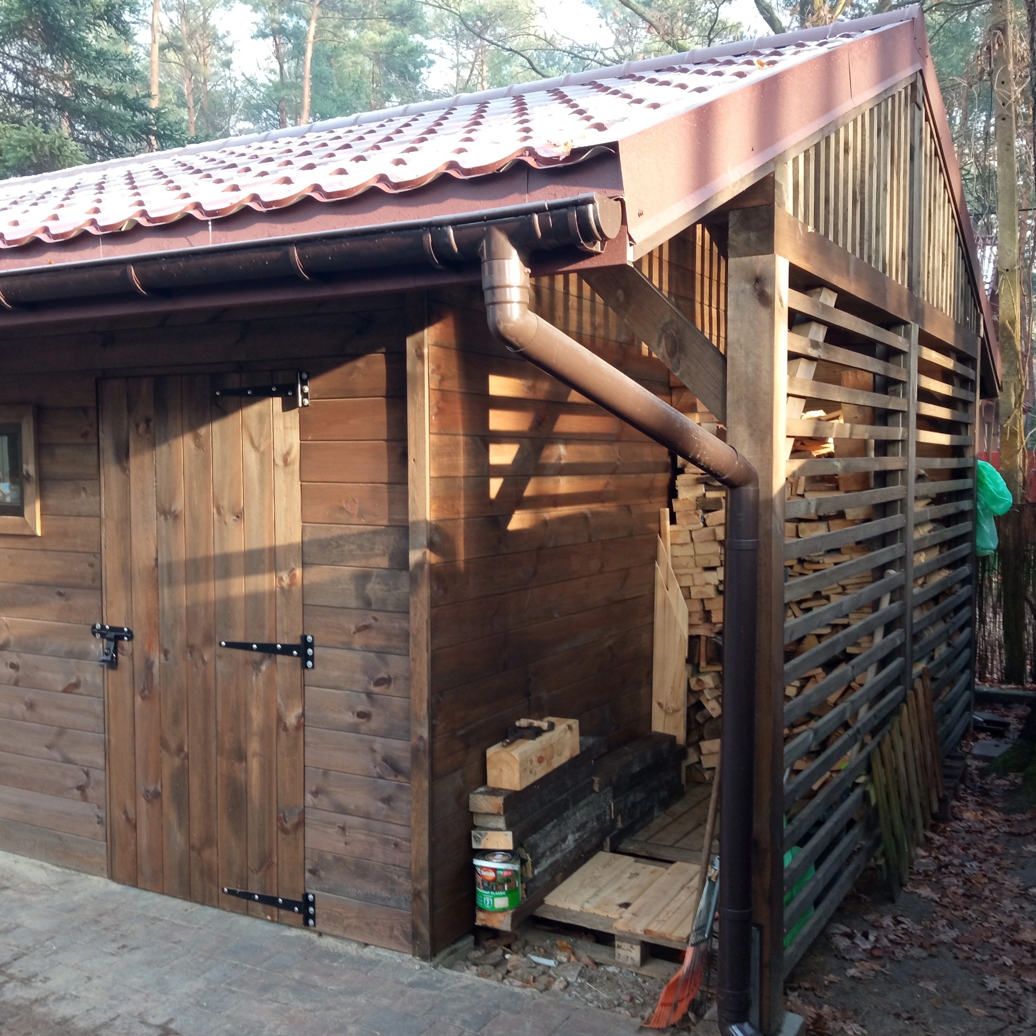 Drewniana konstrukcja z zadaszeniem z blachodachówki, częściowo osłonięta ażurowymi panelami, z widocznym stosem drewna i narzędziami w tle.