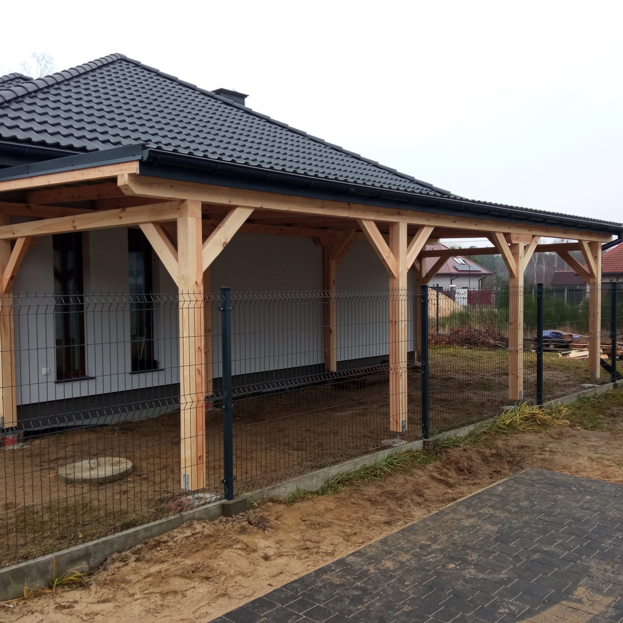 Surowa drewniana konstrukcja z czarnym dachem, prawdopodobnie zadaszenie tarasu lub pergola, w trakcie budowy, widoczna siatka ogrodzeniowa i kostka brukowa.