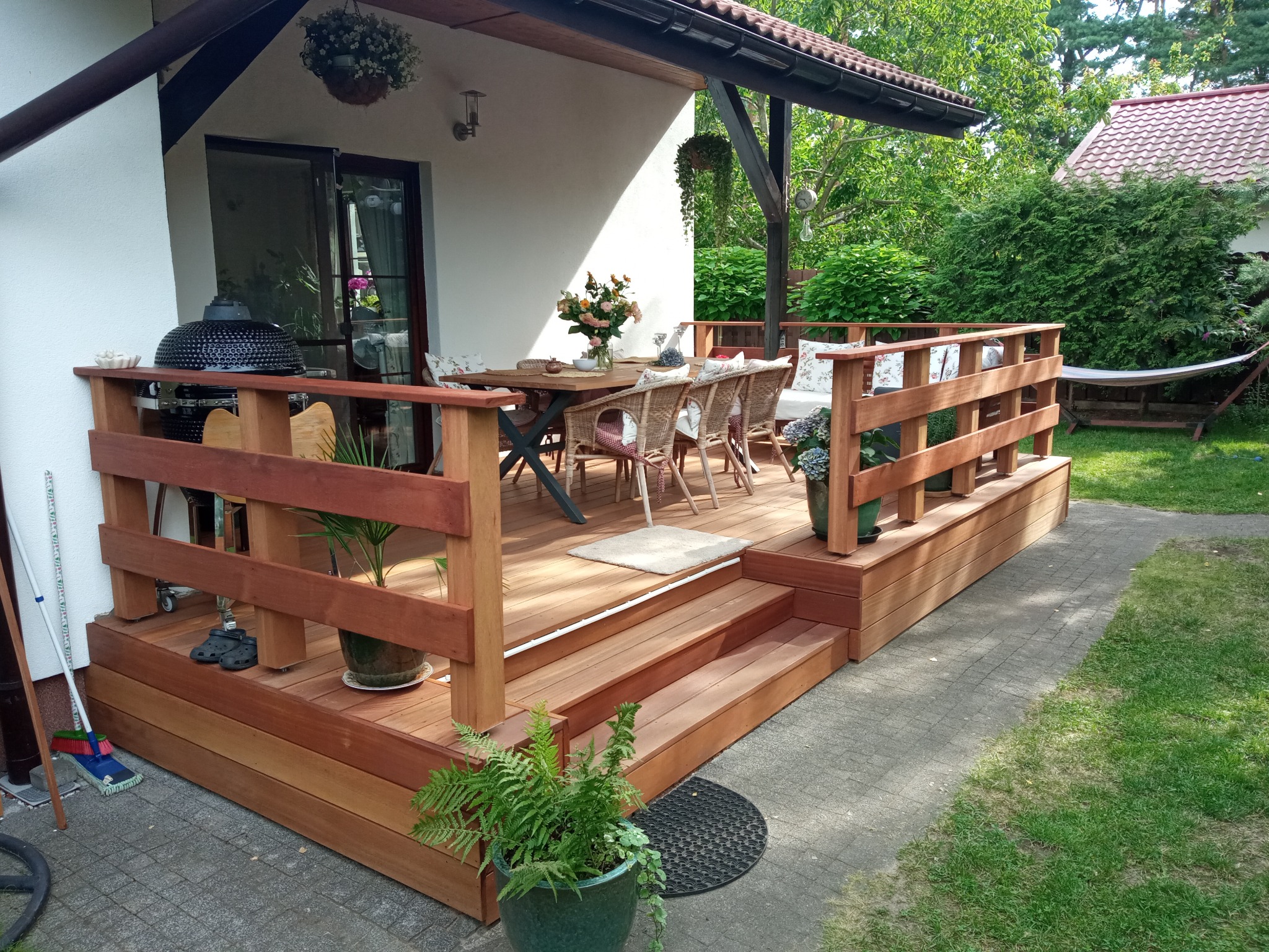 Zadaszony, drewniany taras z balustradą, stołem jadalnym i grillem ceramicznym, otoczony zielenią ogrodu.