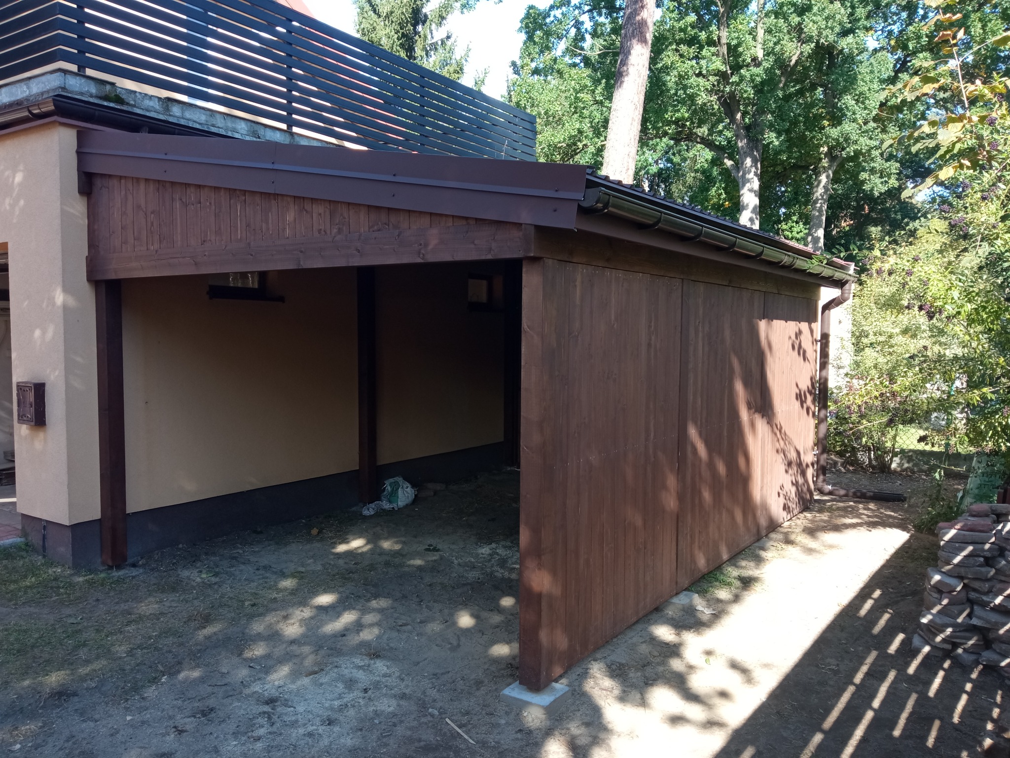 Drewniana konstrukcja z zadaszeniem, prawdopodobnie wiata garażowa, z elementami zadaszenia w kolorze brązowym, widok z zewnątrz, na ziemi widoczne ślady cienia drzew.