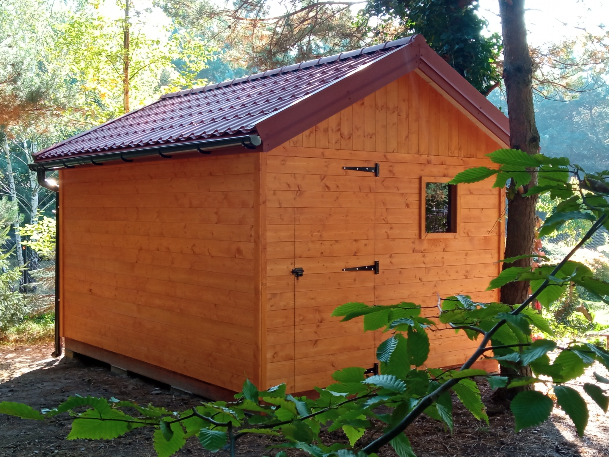 Mały, drewniany budynek z brązowym dachem z blachy trapezowej, otoczony drzewami i zielenią, z widocznymi zawiasami i małym oknem.