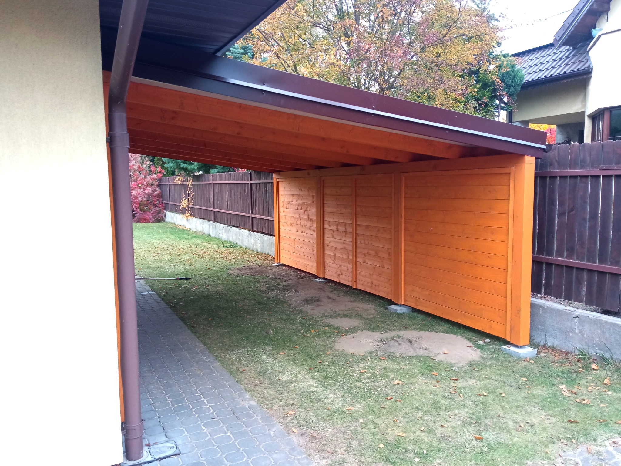 Zadaszone miejsce parkingowe z drewna, pomalowane na pomarańczowo-brązowy kolor, przylegające do budynku, widoczne z zewnątrz na tle jesiennego ogrodu.