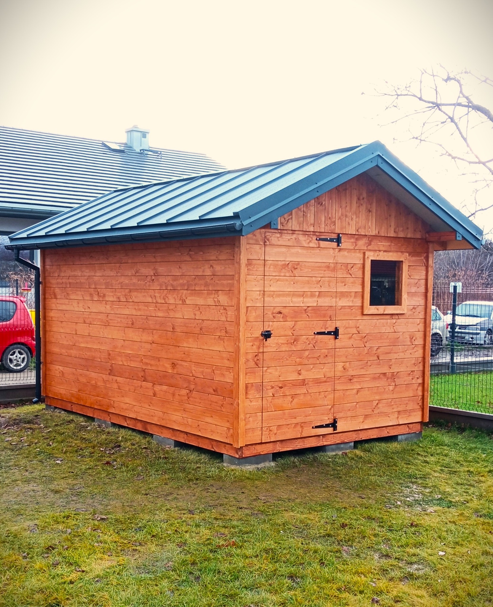 Drewniana altana ogrodowa z oknem i metalowym dachem, umieszczona na betonowych bloczkach, otoczona trawnikiem i ogrodzeniem.
