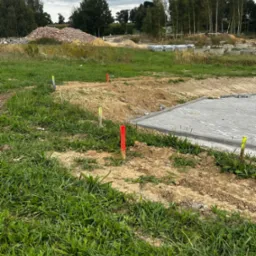 Wytyczanie terenu pod budowę z użyciem palików geodezyjnych oznaczonych kolorowymi taśmami, widoczny fragment ułożonej kostki brukowej i hałda gruzu w tle.