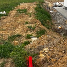 XiGEO.PL - Teren budowy z wytyczonymi punktami geodezyjnymi oznaczonymi czerwonymi słupkami, widoczne wykopy i przygotowanie pod chodnik z kostki brukowej.