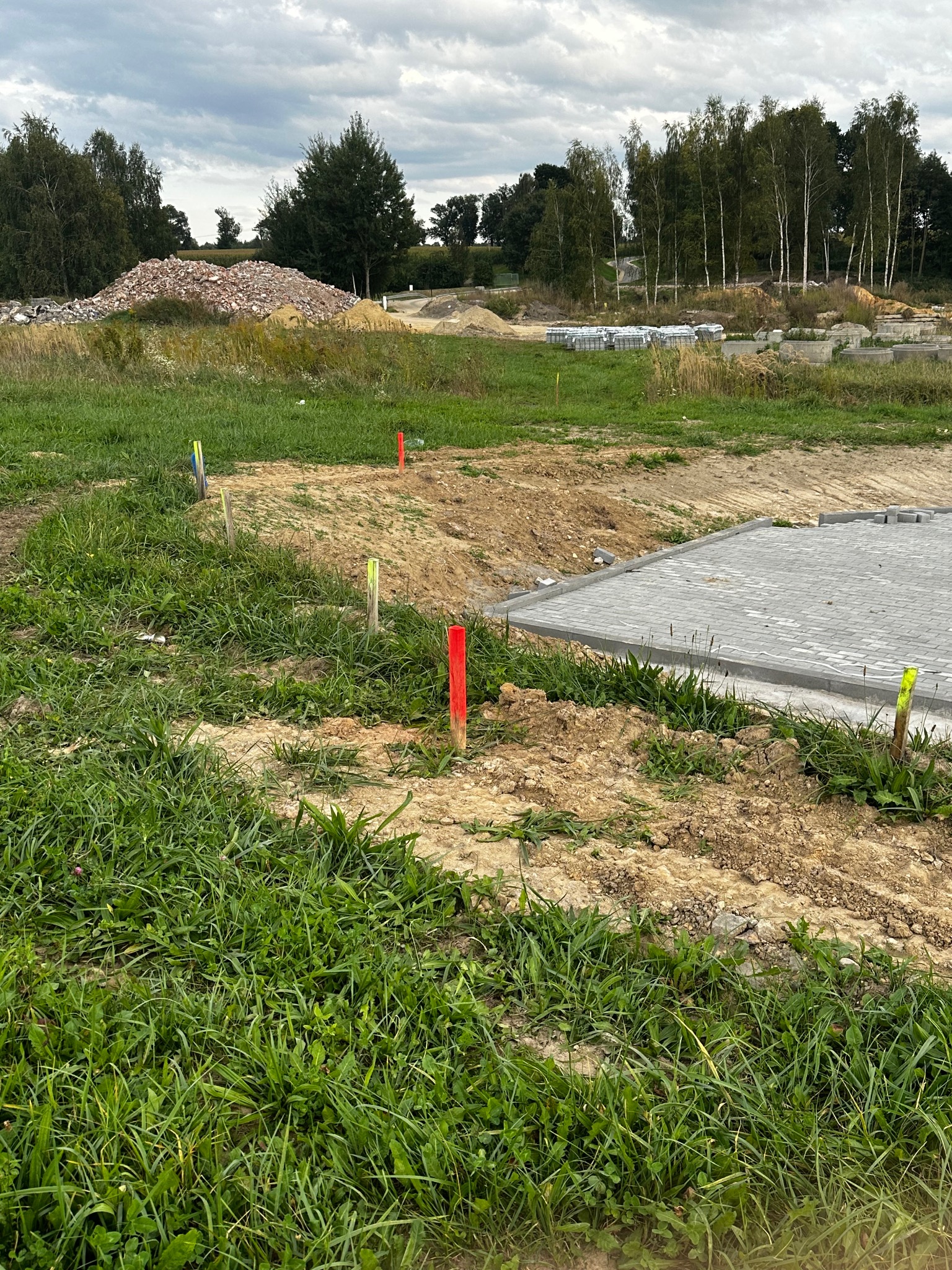 Wytyczanie terenu pod budowę z użyciem palików geodezyjnych oznaczonych kolorowymi taśmami, widoczny fragment ułożonej kostki brukowej i hałda gruzu w tle.