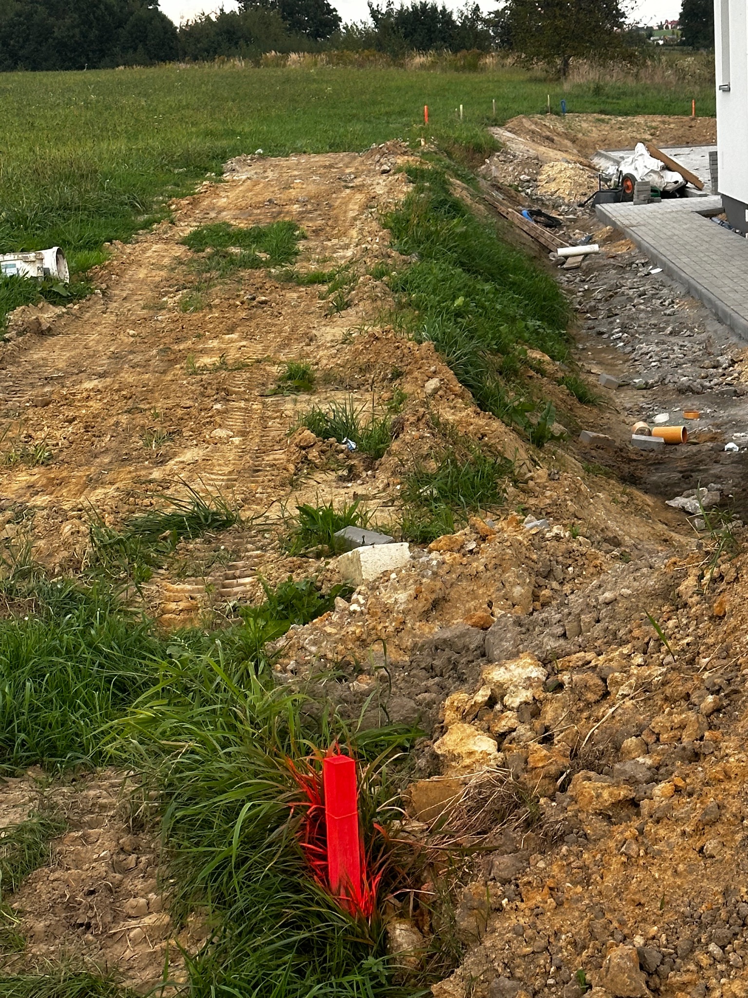 Teren budowy z wytyczonymi punktami geodezyjnymi oznaczonymi czerwonymi słupkami, widoczne wykopy i przygotowanie pod chodnik z kostki brukowej.