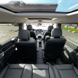 Wnętrze luksusowego busa z czarną skórzaną tapicerką, widok z tyłu, panoramiczny dach, butelki wody w uchwytach, widok na parking i budynek w tle.