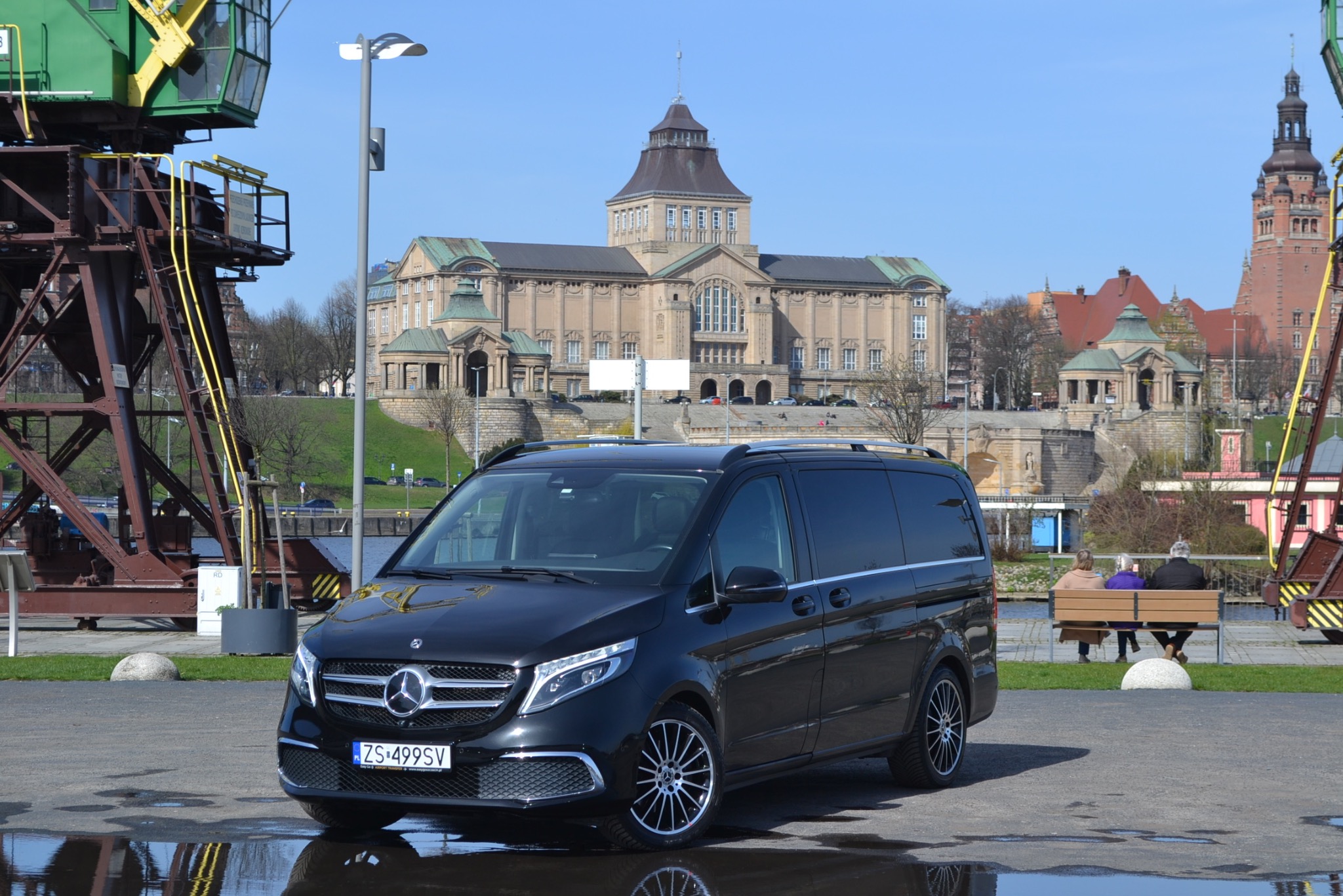 Czarny van Mercedes-Benz V-klasy na tle Muzeum Narodowego w Szczecinie i dźwigu portowego, z odbiciem w kałuży na pierwszym planie.