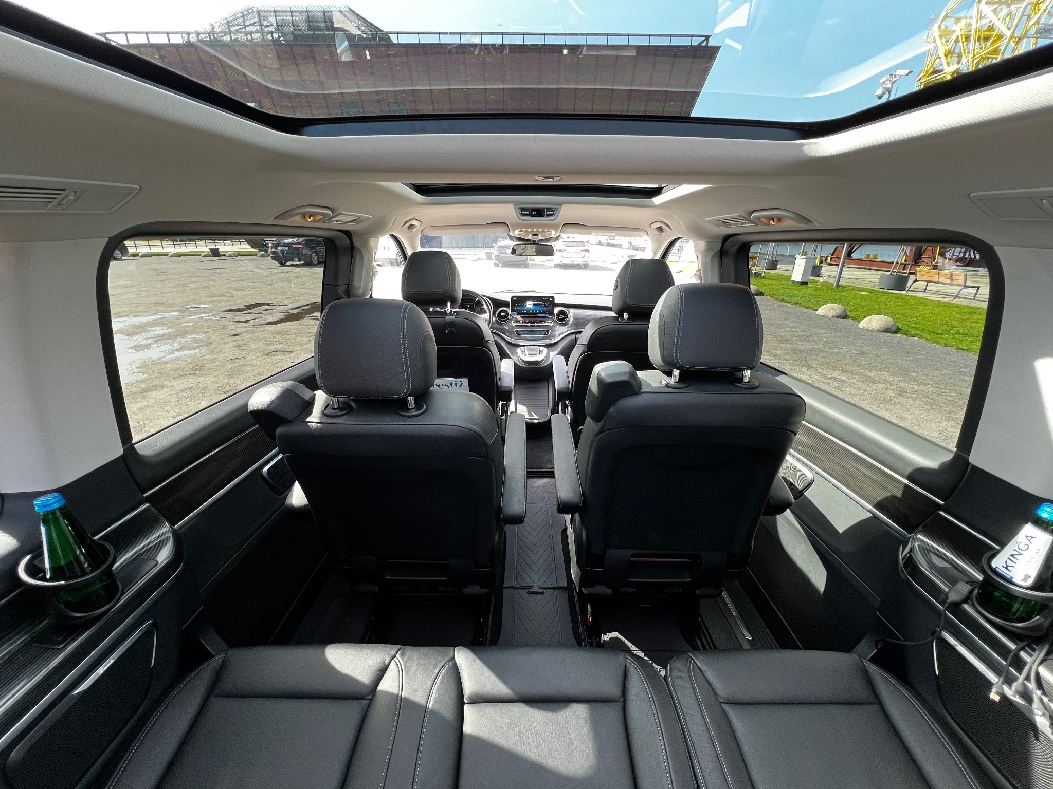 Wnętrze luksusowego busa z czarną skórzaną tapicerką, widok z tyłu, panoramiczny dach, butelki wody w uchwytach, widok na parking i budynek w tle.