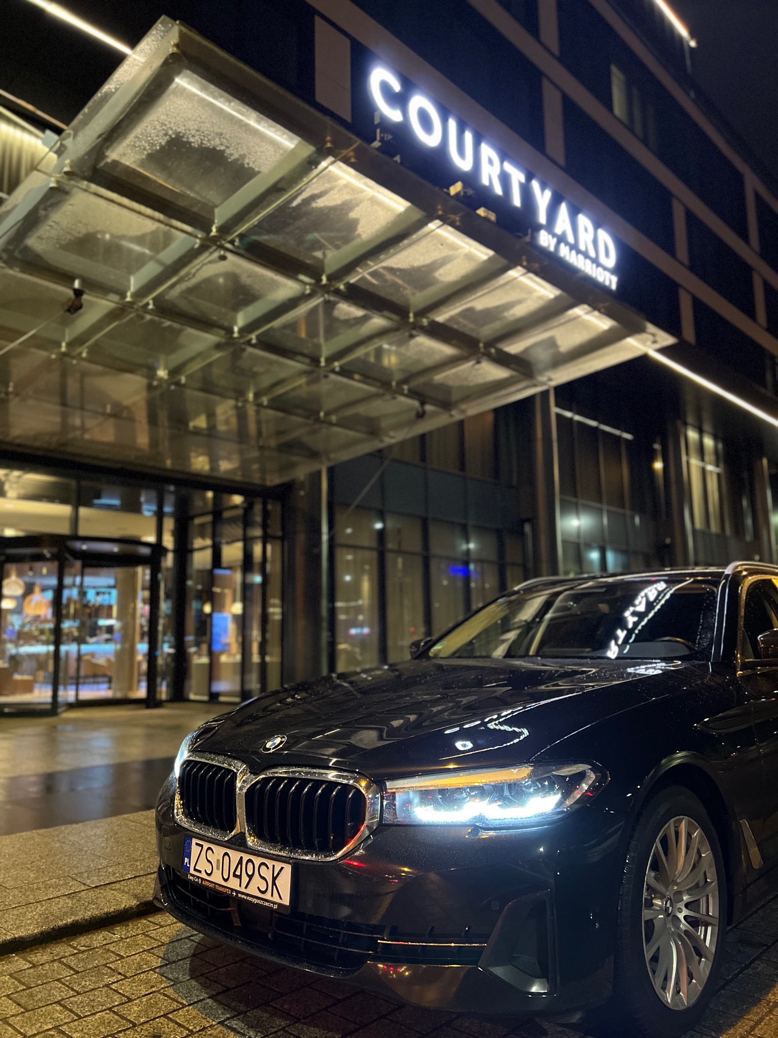 Czarna limuzyna BMW zaparkowana przed hotelem Courtyard by Marriott nocą, widoczna tablica rejestracyjna ZS 049SK.