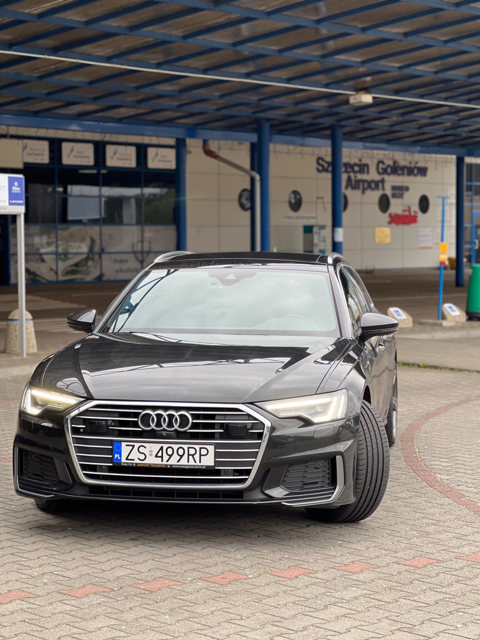Czarna limuzyna Audi A6 kombi zaparkowana przed lotniskiem Szczecin-Goleniów, widoczna tablica rejestracyjna z reklamą transferu lotniskowego.