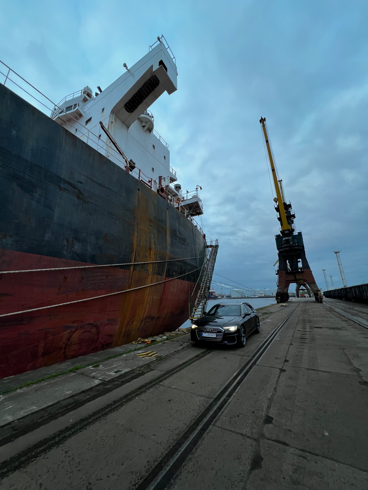 Czarny samochód osobowy Audi na tle nabrzeża portowego z dźwigiem i dużym statkiem towarowym w pochmurny dzień.