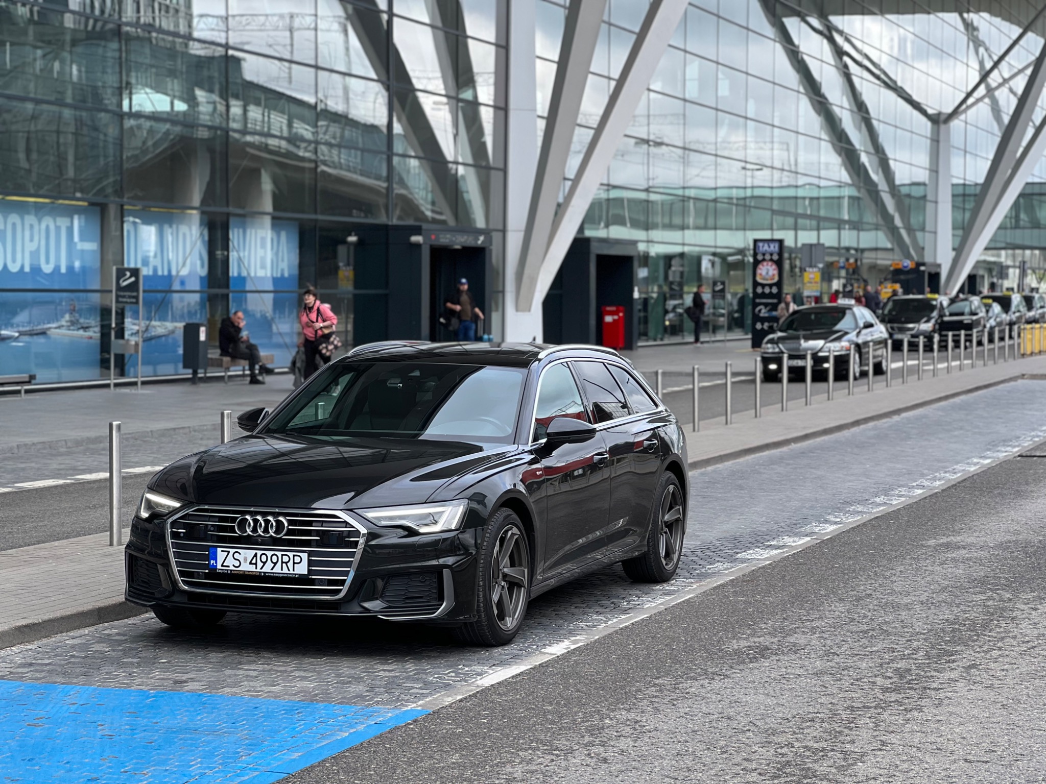 Czarny samochód Audi kombi z numerem rejestracyjnym ZS 499RP zaparkowany przed nowoczesnym terminalem lotniska, w tle rząd podobnych pojazdów i osoby oczekujące.