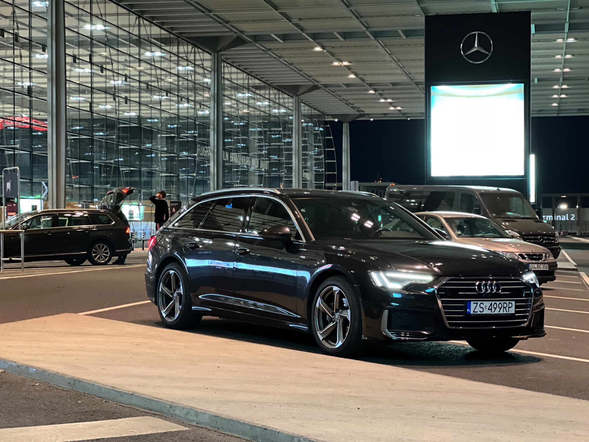 Czarny samochód kombi Audi A6 zaparkowany przed lotniskiem Berlin Brandenburg Willy Brandt w nocy, z otwartym bagażnikiem innego pojazdu w tle.