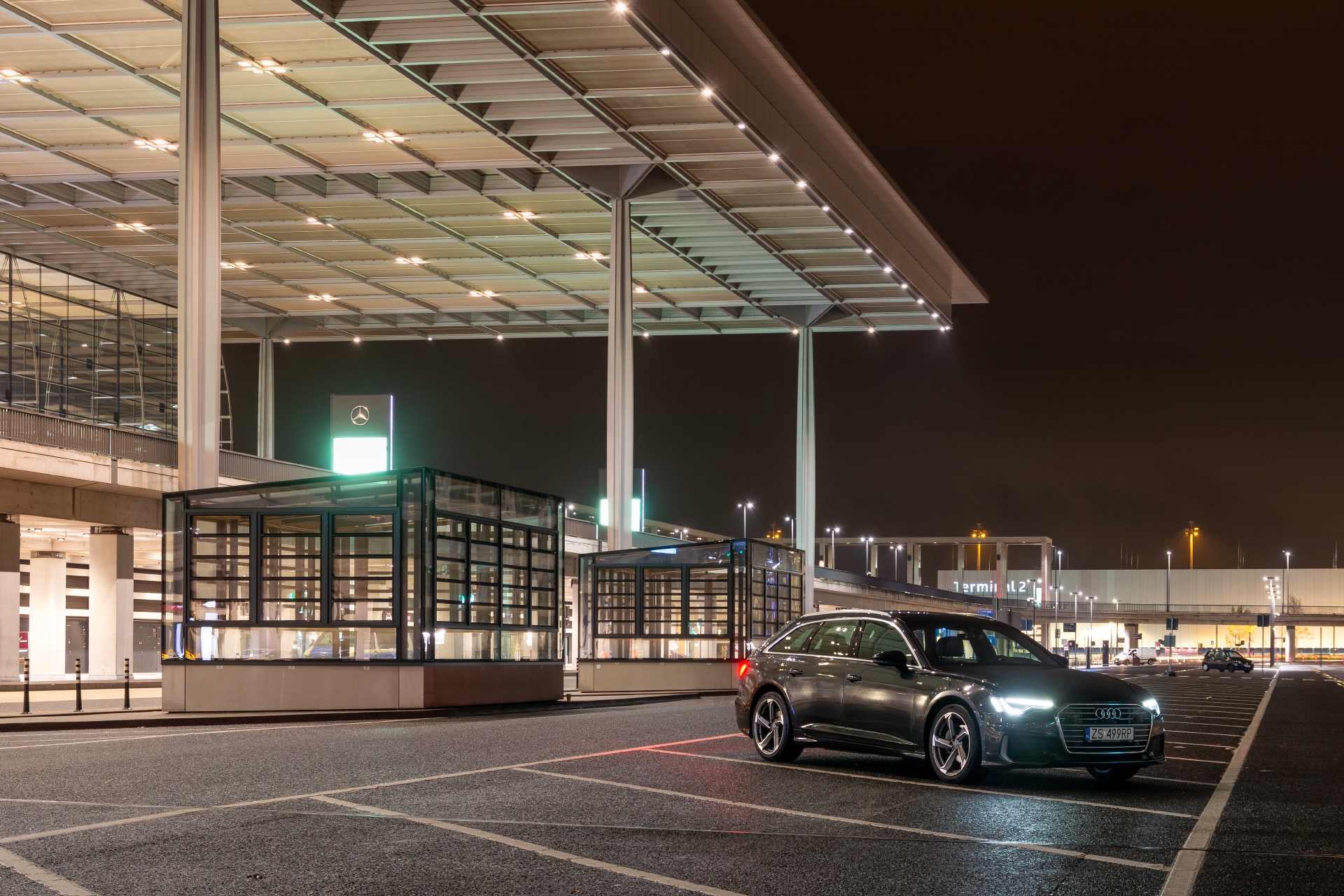 Grafitowe Audi A6 Avant na parkingu lotniska nocą, z widocznym oświetleniem terminalu i znakiem Mercedesa w tle.