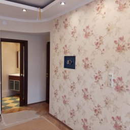 Roman Andrushchenko Akwarystyka Profesjonalna - Ściana z kwiatową tapetą w Ostrowie Wielkopolskim, z widoczną puszką elektryczną i gniazdkiem. W tle widoczne drzwi do łazienki z wykończoną podłogą w szachownicę.