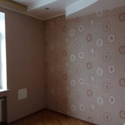 Roman Andrushchenko Akwarystyka Profesjonalna - Pokój po remoncie: ściany pokryte tapetą w geometryczne wzory, drewniana podłoga, grzejnik pod oknem, mała klapka inspekcyjna w ścianie.