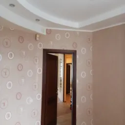 Pokój po remoncie: ściany w beżowej tapecie z motywem kół i liter, drewniana podłoga, otwarte drzwi do kolejnego pomieszczenia, sufit z ozdobnym oświetleniem.