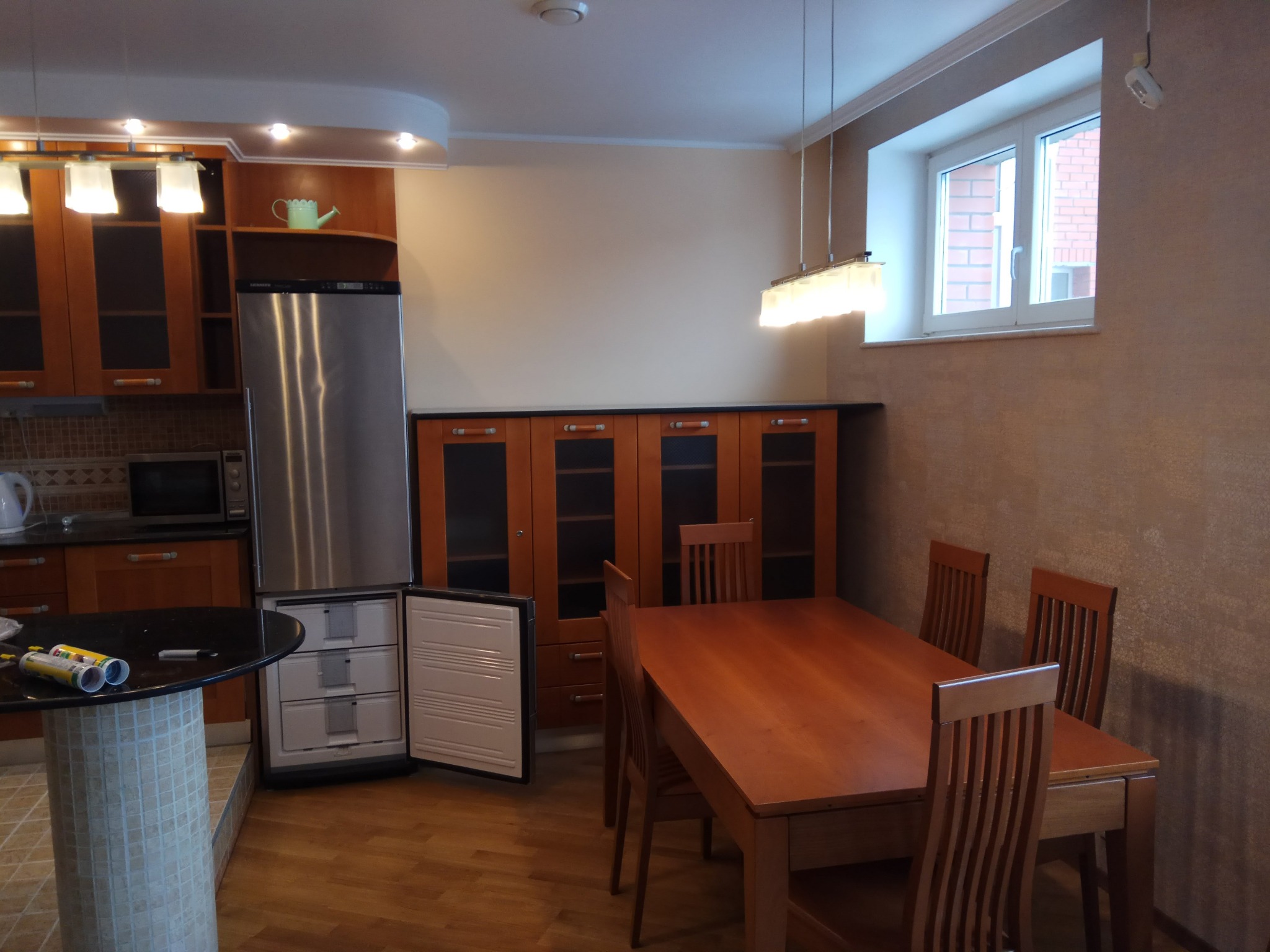 Umeblowana kuchnia z otwartą szufladą zamrażarki, widoczne narzędzia na blacie stołu, drewniany stół jadalny z krzesłami, brązowe szafki i okno z widokiem na ceglany budynek.