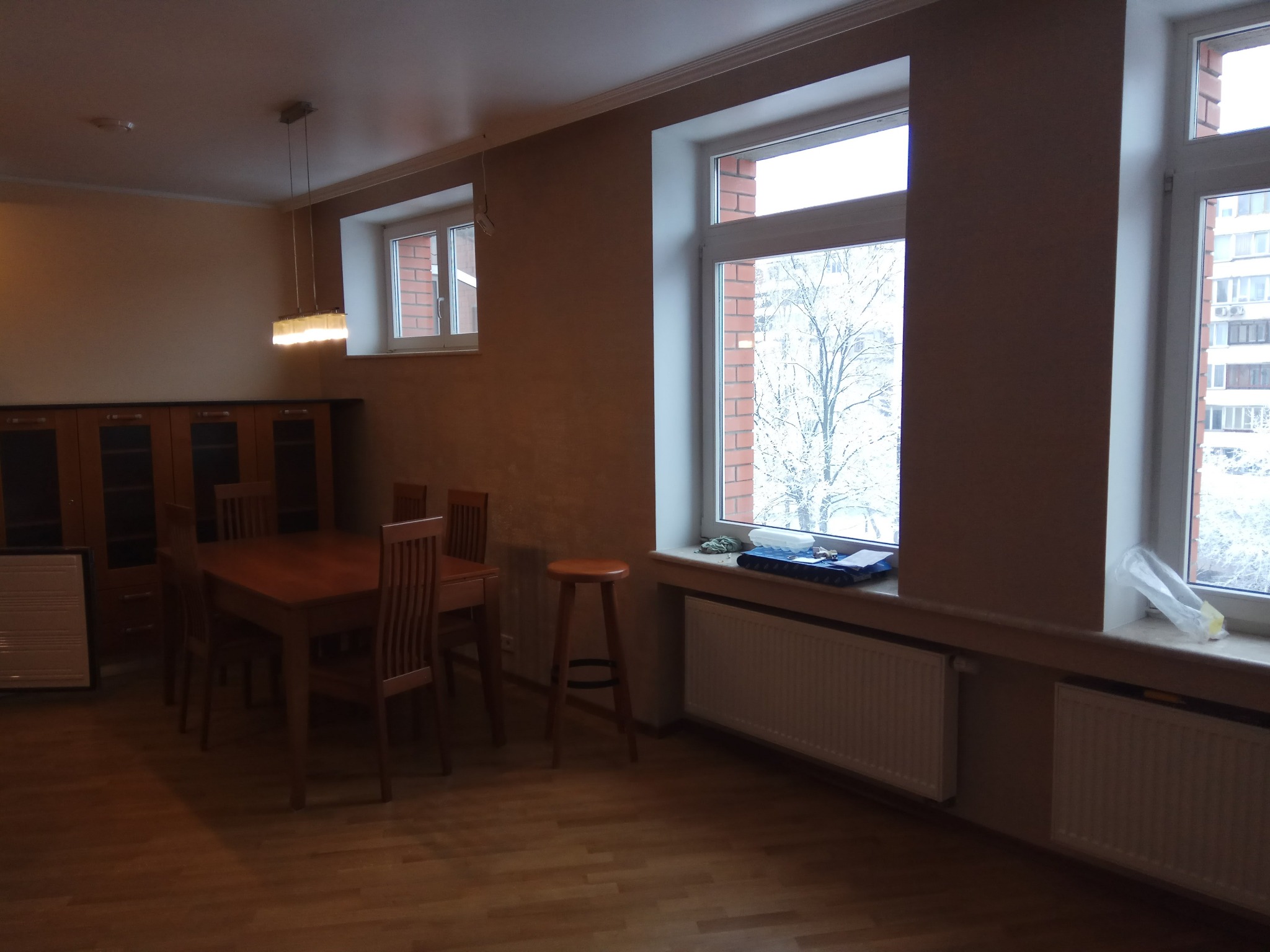 Jasne wnętrze jadalni z drewnianym stołem i krzesłami, widoczne grzejniki pod oknami i wisząca lampa sufitowa.