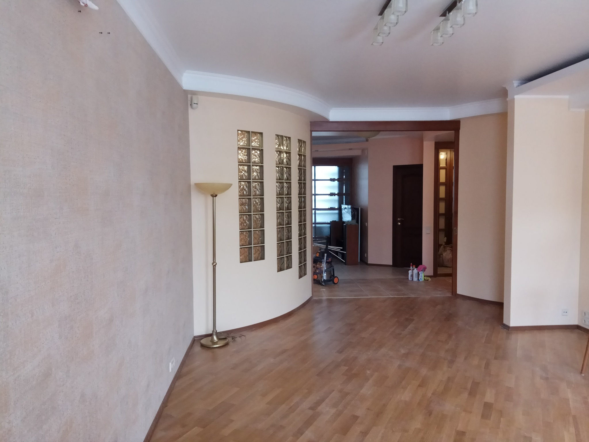 Przestronne wnętrze mieszkania z drewnianą podłogą, widoczną ścianą z luksferami i lampą podłogową, w tle widoczny sprzęt do sprzątania.