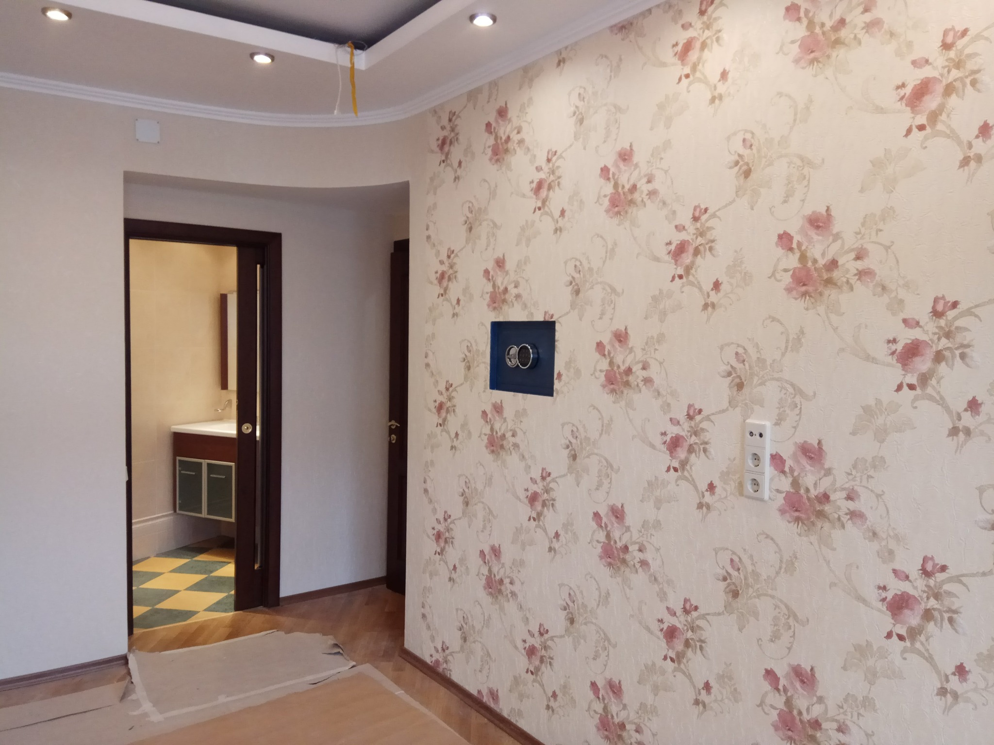 Ściana z kwiatową tapetą w Ostrowie Wielkopolskim, z widoczną puszką elektryczną i gniazdkiem. W tle widoczne drzwi do łazienki z wykończoną podłogą w szachownicę.