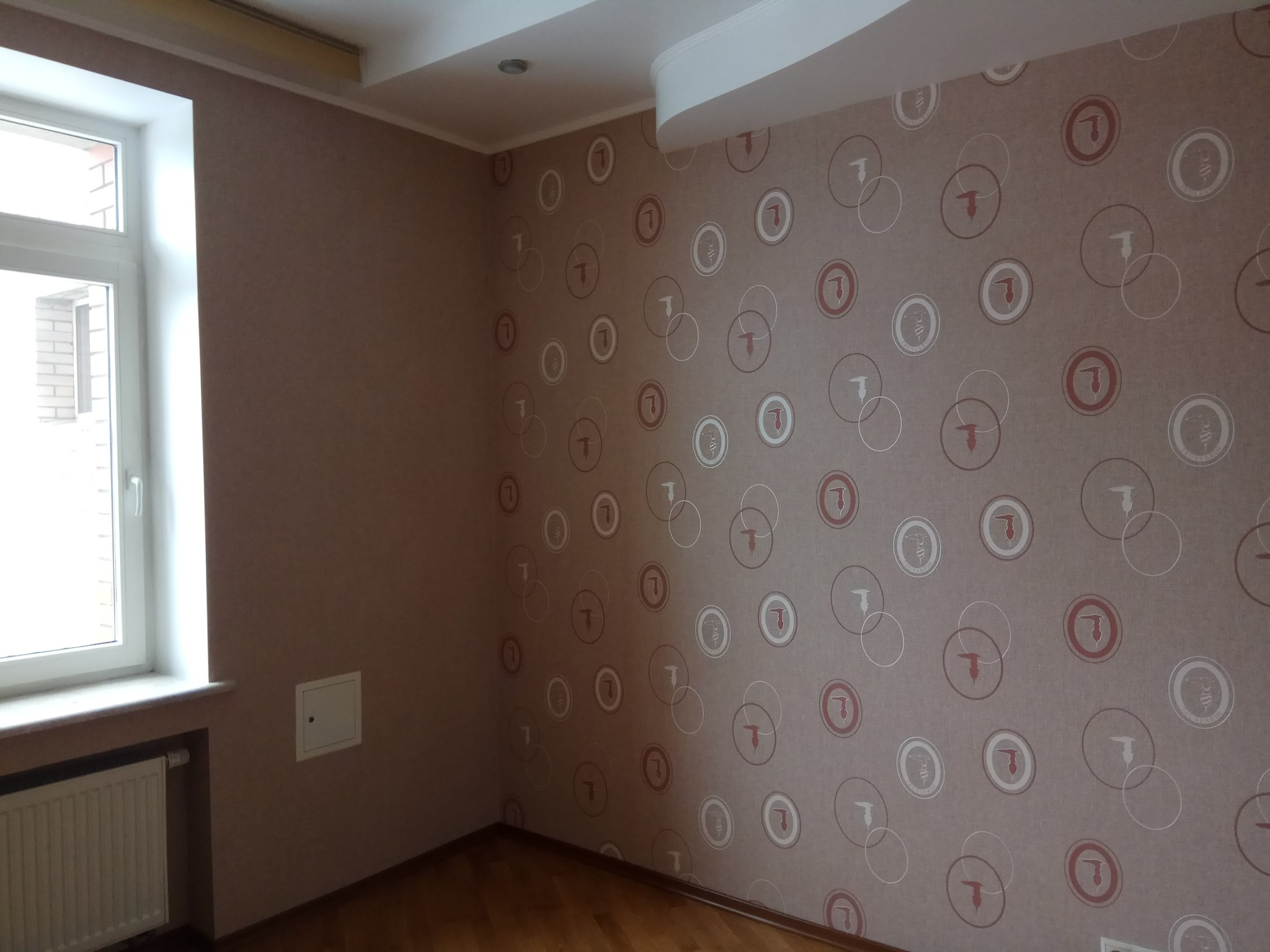 Pokój po remoncie: ściany pokryte tapetą w geometryczne wzory, drewniana podłoga, grzejnik pod oknem, mała klapka inspekcyjna w ścianie.