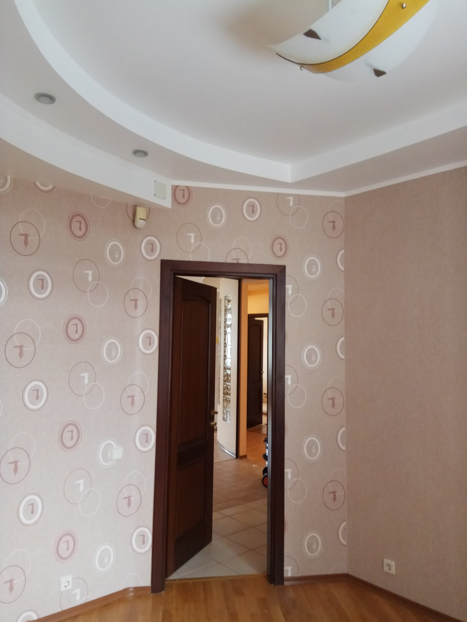 Pokój po remoncie: ściany w beżowej tapecie z motywem kół i liter, drewniana podłoga, otwarte drzwi do kolejnego pomieszczenia, sufit z ozdobnym oświetleniem.