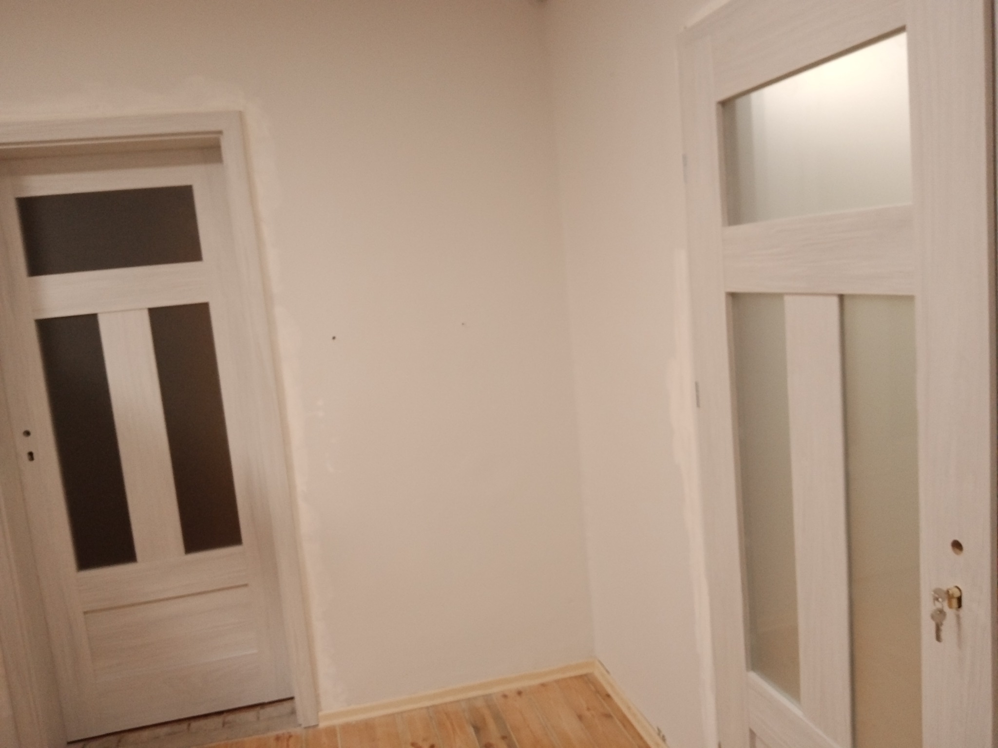 Dwa skrzydła drzwiowe z przeszkleniami w jasnej tonacji, jedno otwarte, drugie zamknięte z kluczem w zamku, widoczne na tle białych ścian i drewnianej podłogi.