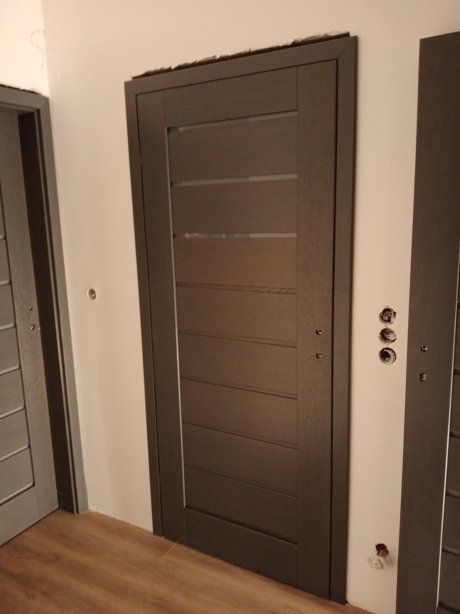 Ciemnoszare drzwi wewnętrzne z poziomymi przeszkleniami, widoczne otwory w ścianie po instalacji elektrycznej, podłoga drewnopodobna.