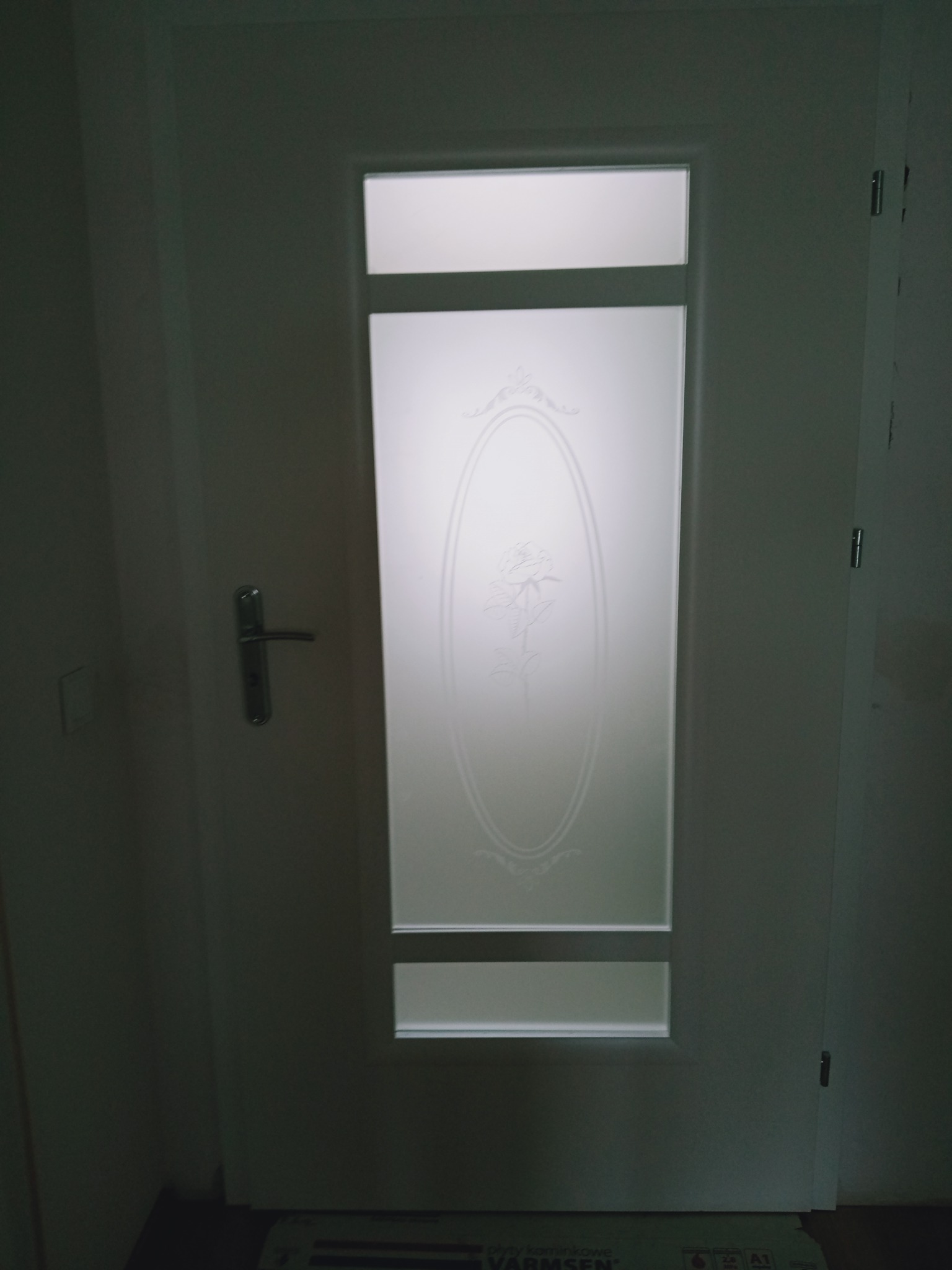 Białe drzwi wewnętrzne z matową szybą zdobioną motywem róży w owalnej ramie, widoczne zawiasy i klamka.