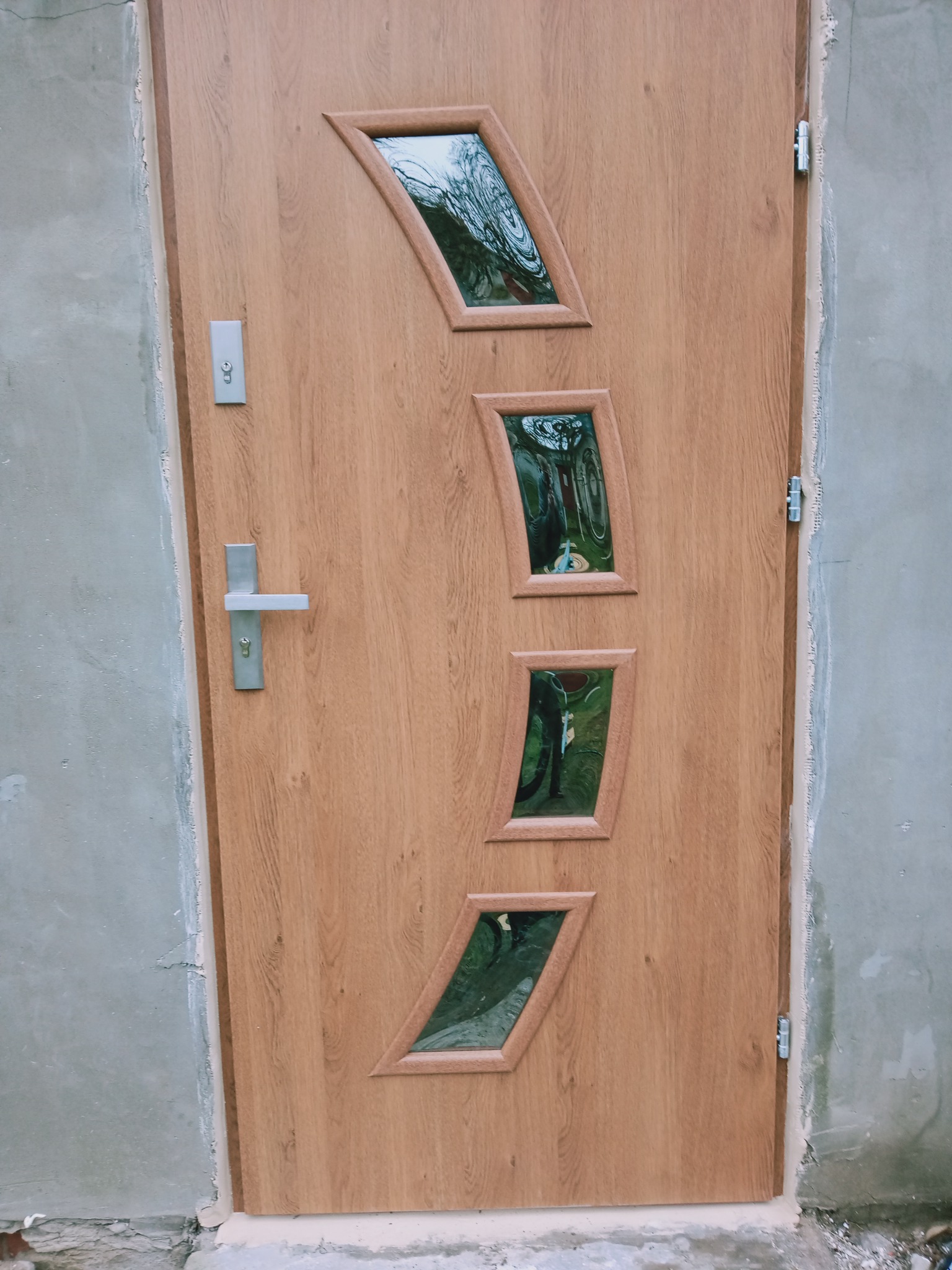 Jasnobrązowe drzwi zewnętrzne z czterema ukośnymi szybkami o nieregularnych kształtach, srebrna klamka, widoczne ślady montażu na ścianie.