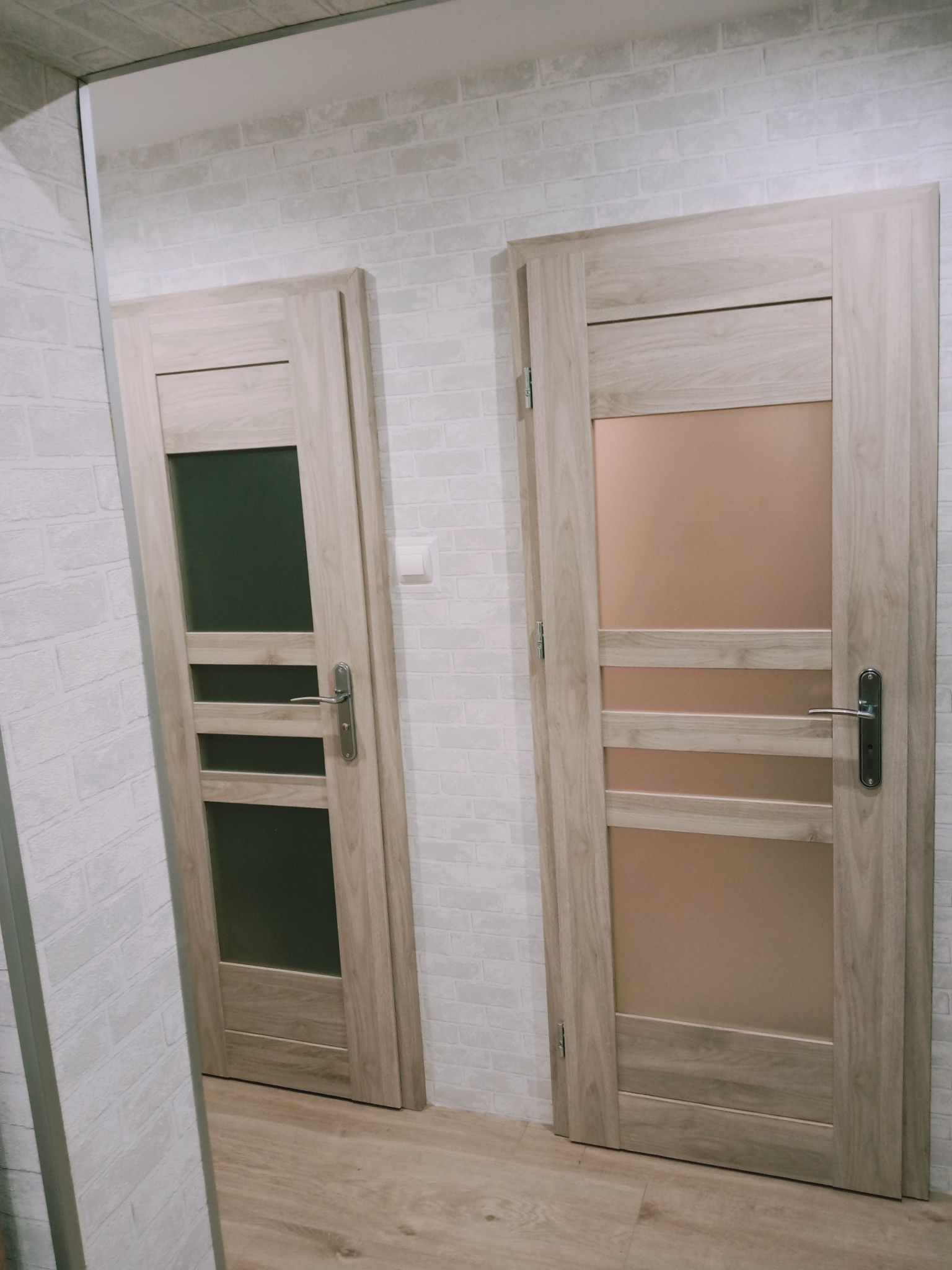 Dwie drewniane ościeżnice z szybami w odcieniach szarości i brązu, zamontowane w ścianie z białej cegły, widoczne z perspektywy pod lekkim kątem.