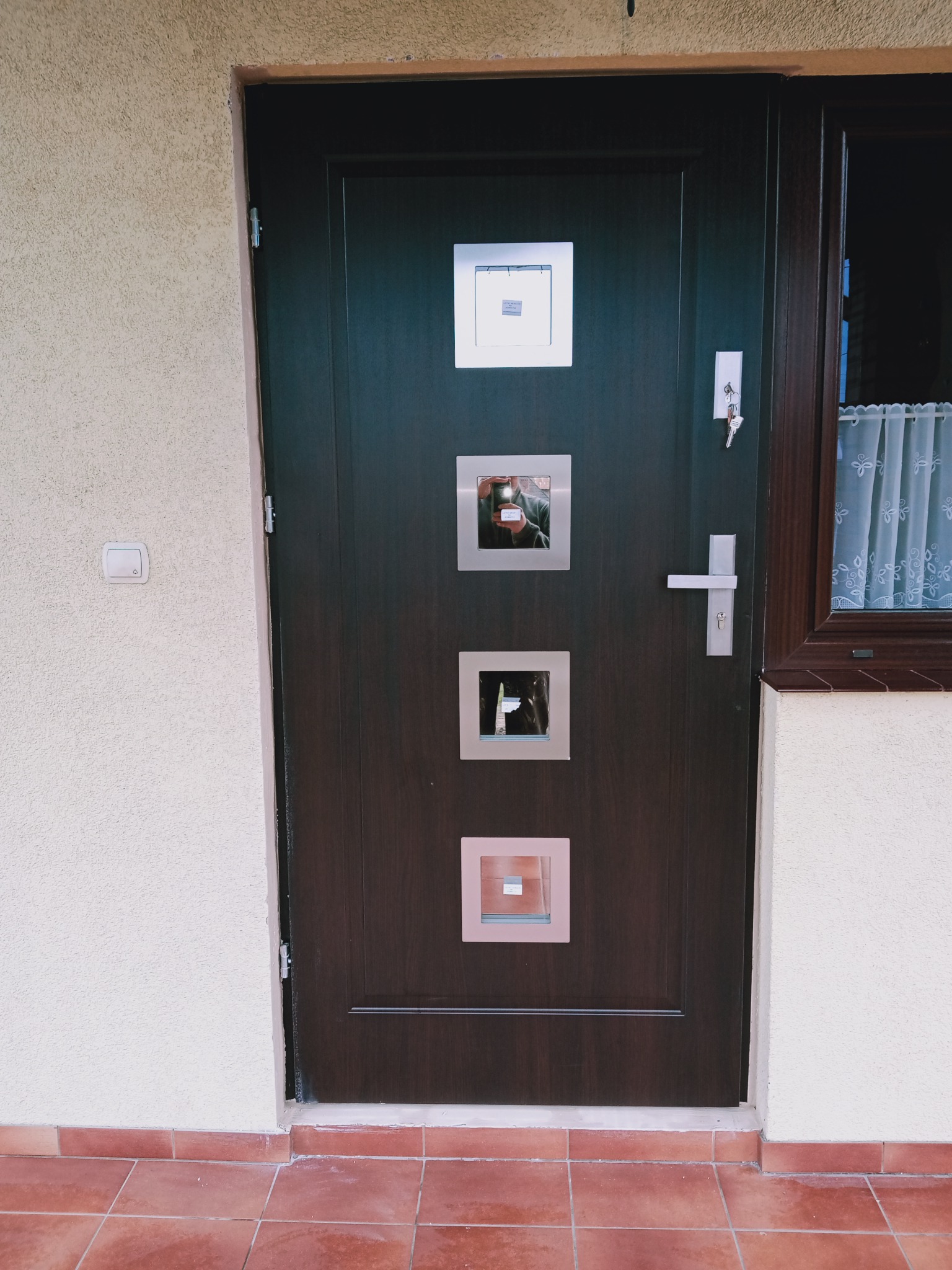 Ciemnobrązowe drzwi wejściowe z czterema kwadratowymi, ozdobnymi wstawkami, zamontowane w jasnym budynku z terakotową podłogą.