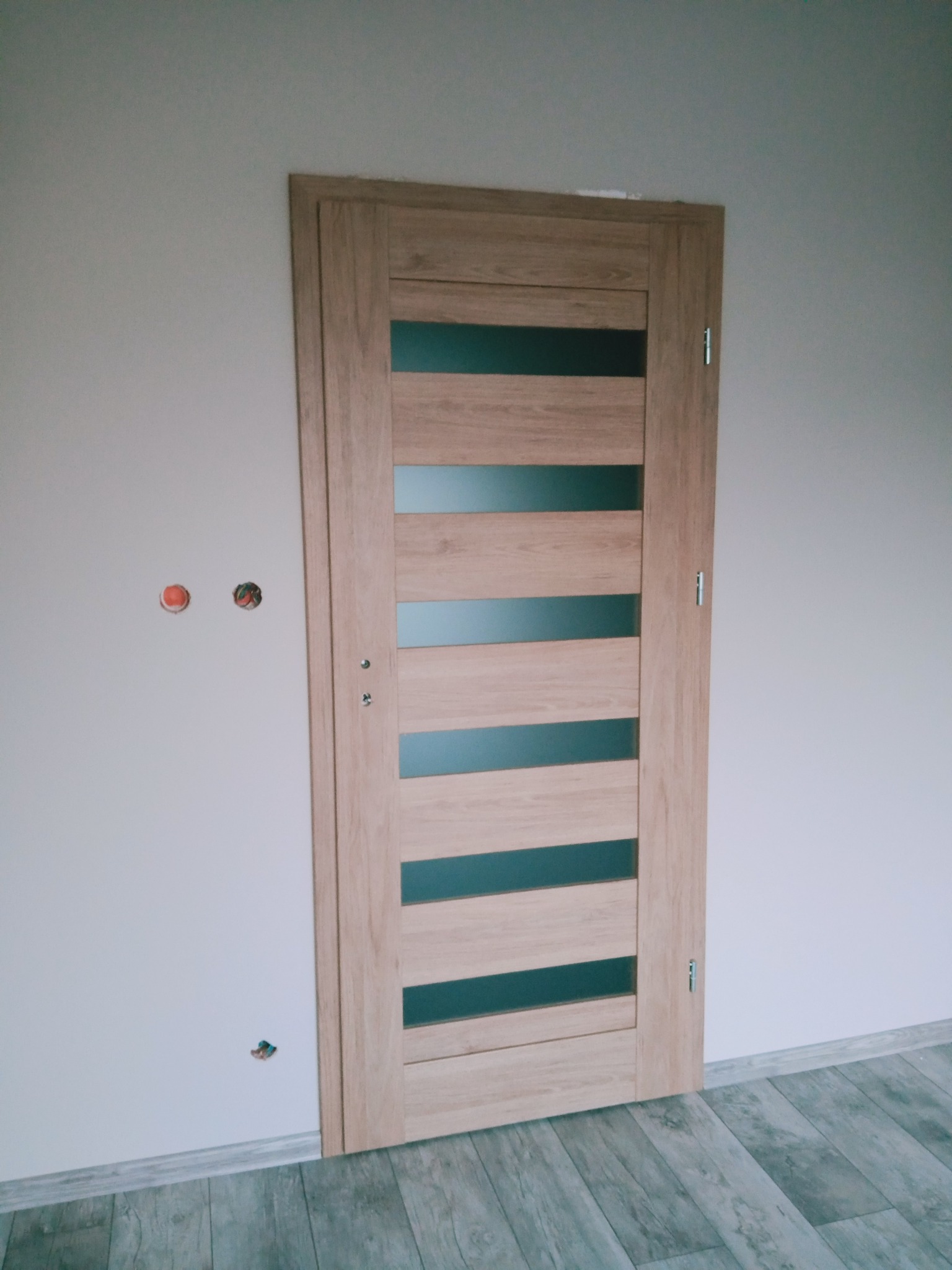 Jasne, drewniane drzwi wewnętrzne z poziomymi, matowymi przeszkleniami, zamontowane w białej ścianie z widocznymi otworami po instalacji elektrycznej i drewnopodobną podłogą.