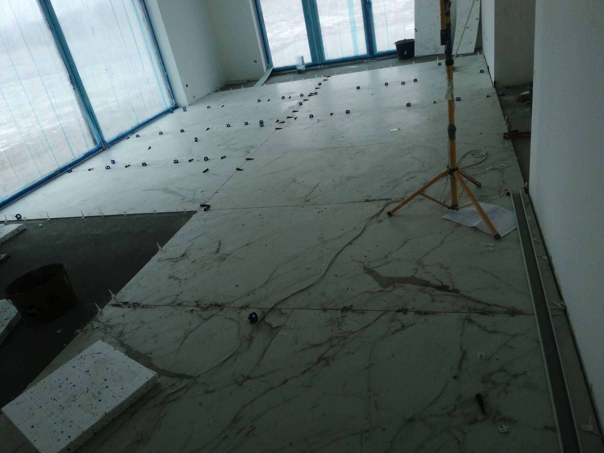 Układanie płytek podłogowych imitujących marmur w jasnym pomieszczeniu z zabezpieczonymi oknami, widoczne kliny poziomujące i lampa robocza.
