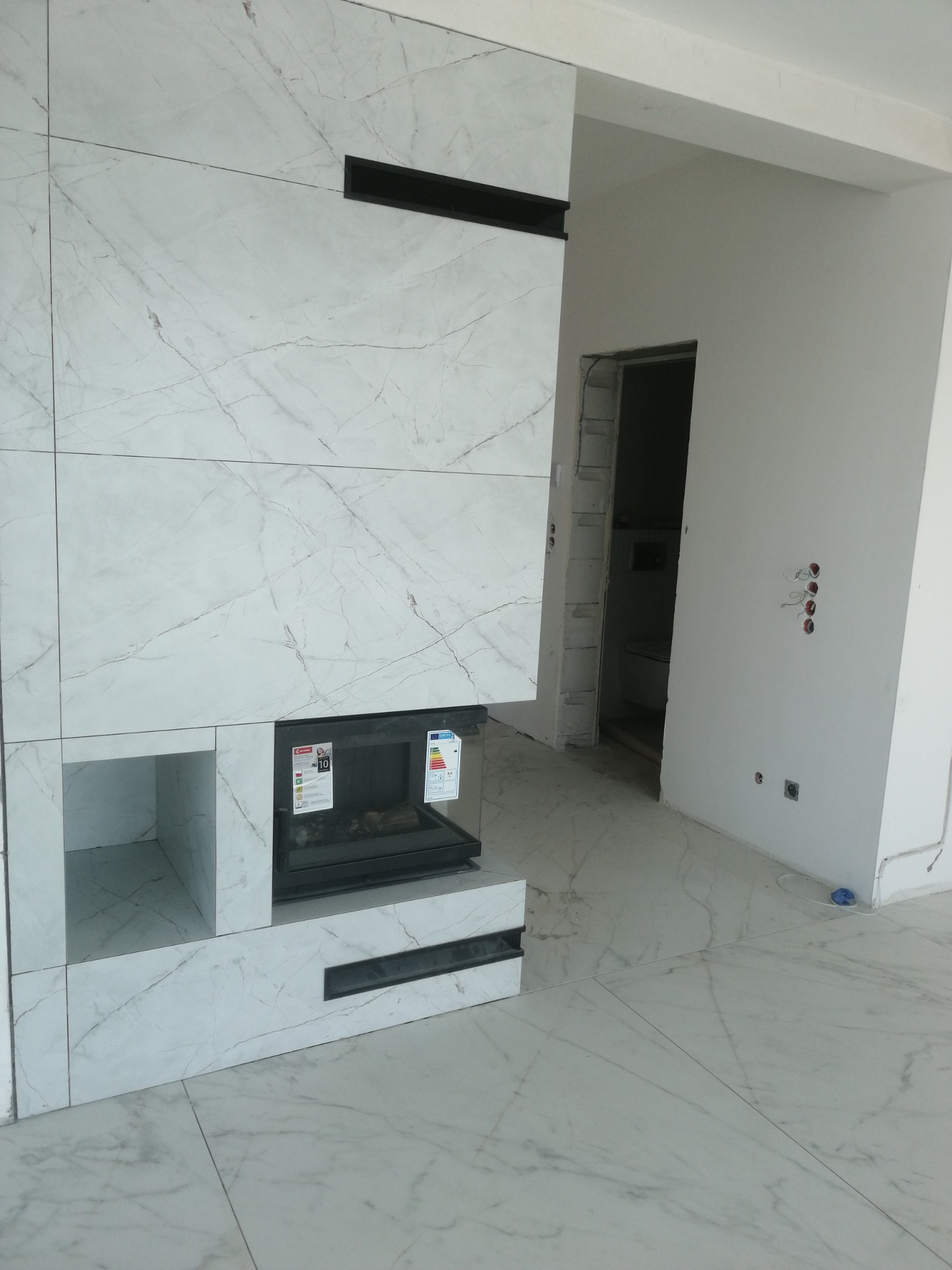 Obudowa kominka z białego marmuru z czarnymi akcentami, widok na wnętrze w trakcie wykańczania, z widocznymi gniazdkami elektrycznymi i otwartym przejściem do innego pomieszczenia.