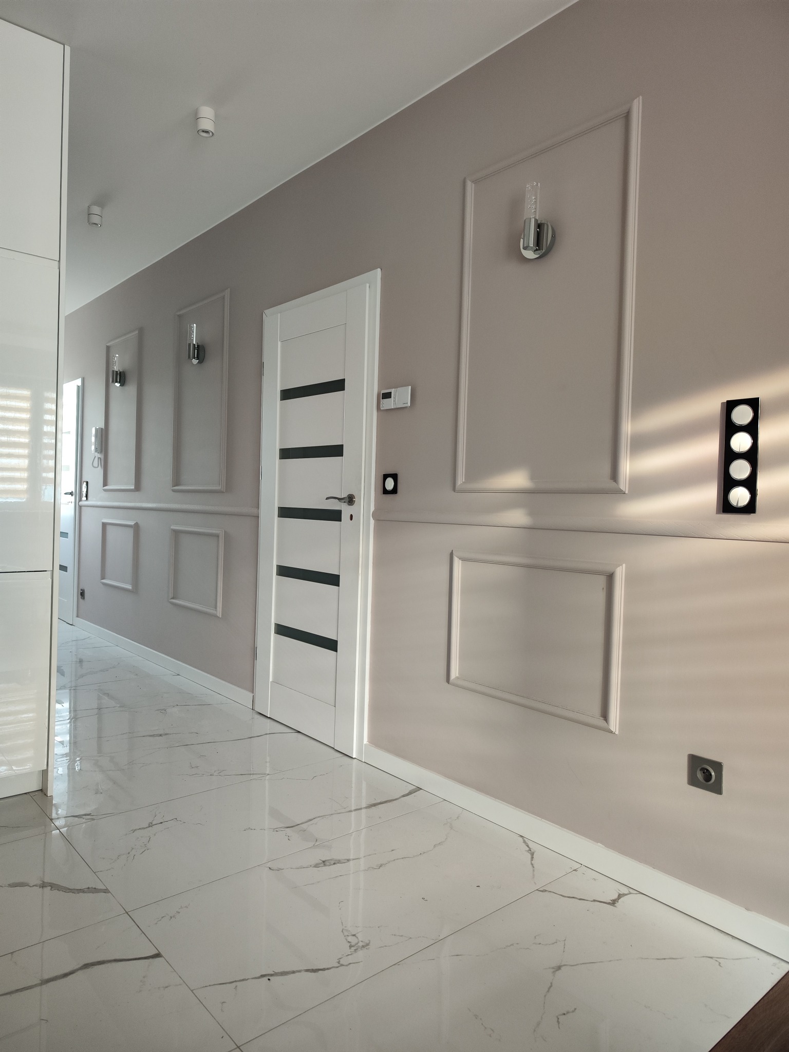 Korytarz w odcieniach beżu z dekoracyjnymi panelami ściennymi, kinkietami, białymi drzwiami z czarnymi paskami i błyszczącą marmurową podłogą.