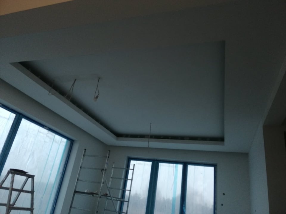 Wykończony sufit podwieszany z wnęką na oświetlenie, widoczne kable, w tle okna zabezpieczone folią i drabiny malarskie.