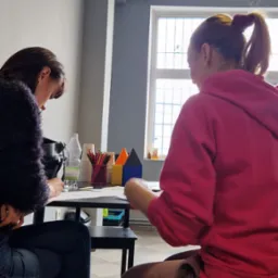 Dwie osoby siedzą przy stole, rysując. Na stole kolorowe kredki, woda w butelce, kartki papieru i geometryczne bryły.