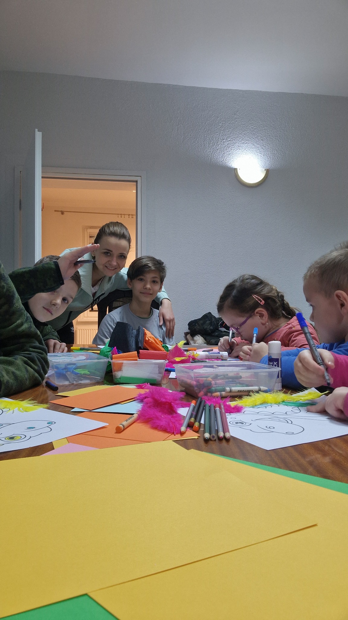 Grupa dzieci i dorosły siedzą przy stole, rysując i kolorując obrazki, na stole kolorowe kartki, kredki, flamastry i piórka.