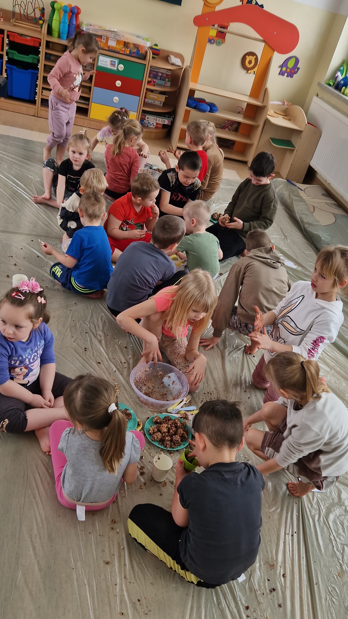 Grupa dzieci siedzi na podłodze, tworząc kulki z miski z brązową masą. Podłoga zabezpieczona folią, w tle regały z zabawkami i dekoracjami.