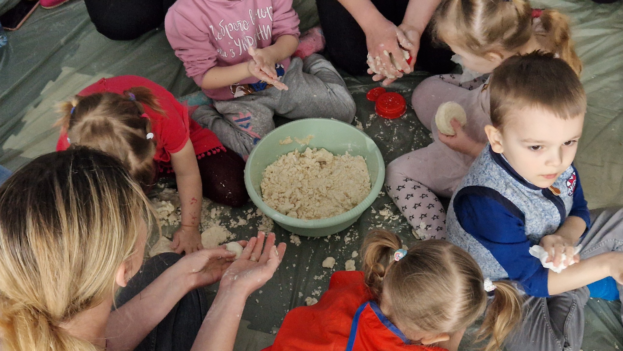 Grupa dzieci siedząca w kręgu wokół miski z ciastem, formująca małe kulki i kształty, na tle zielonej, lekko zabrudzonej powierzchni.