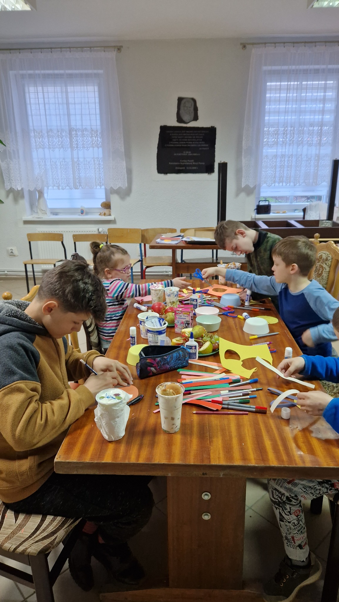 Grupa dzieci przy drewnianym stole podczas zajęć plastycznych: wycinanie i klejenie z kolorowego papieru, przybory plastyczne, owoce i napoje na stole, okno z firanką w tle.