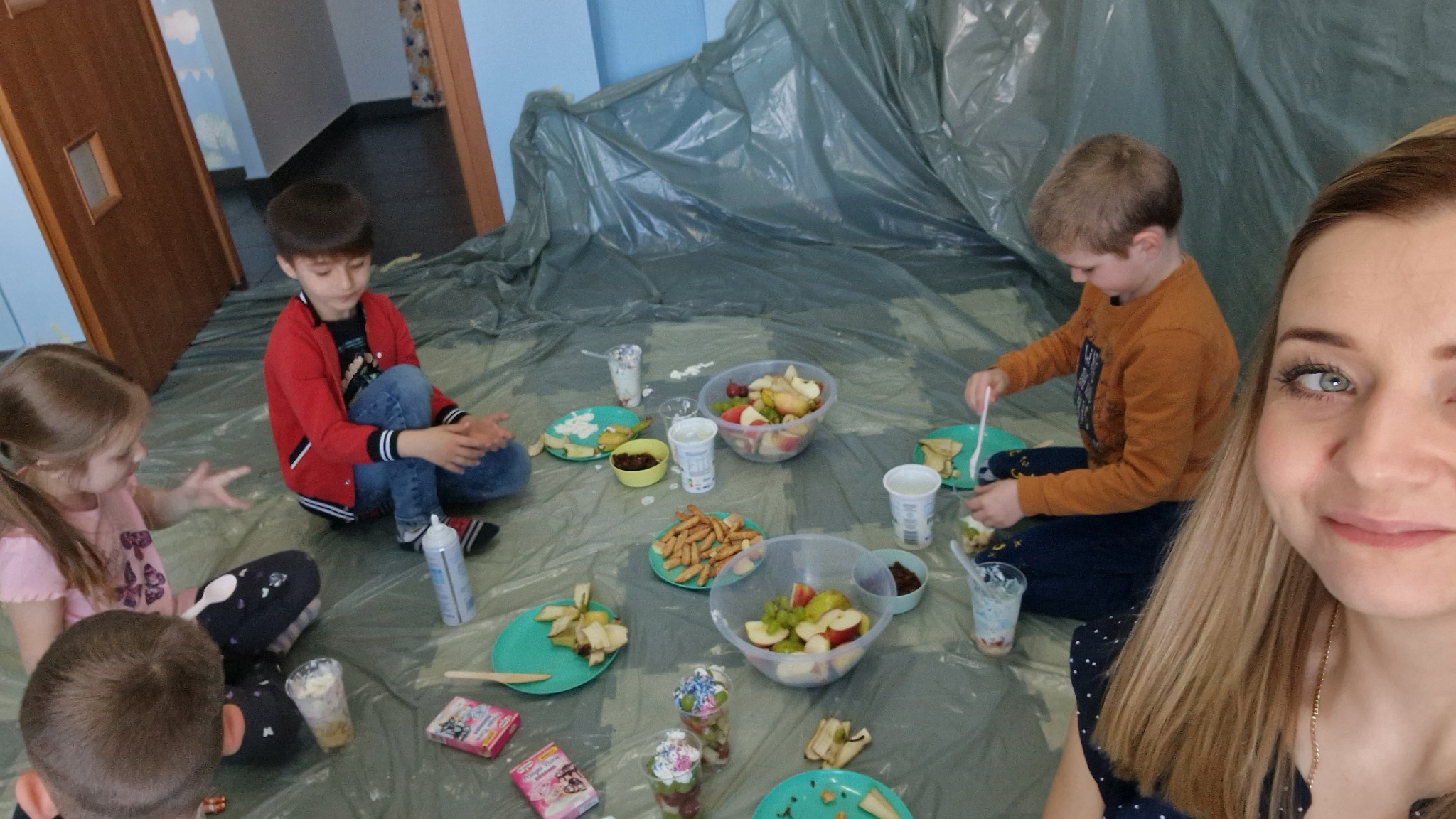 Dzieci siedzą na folii ochronnej, jedząc owoce i przekąski podczas imprezy urodzinowej, selfie z uśmiechniętą kobietą w prawym dolnym rogu kadru.