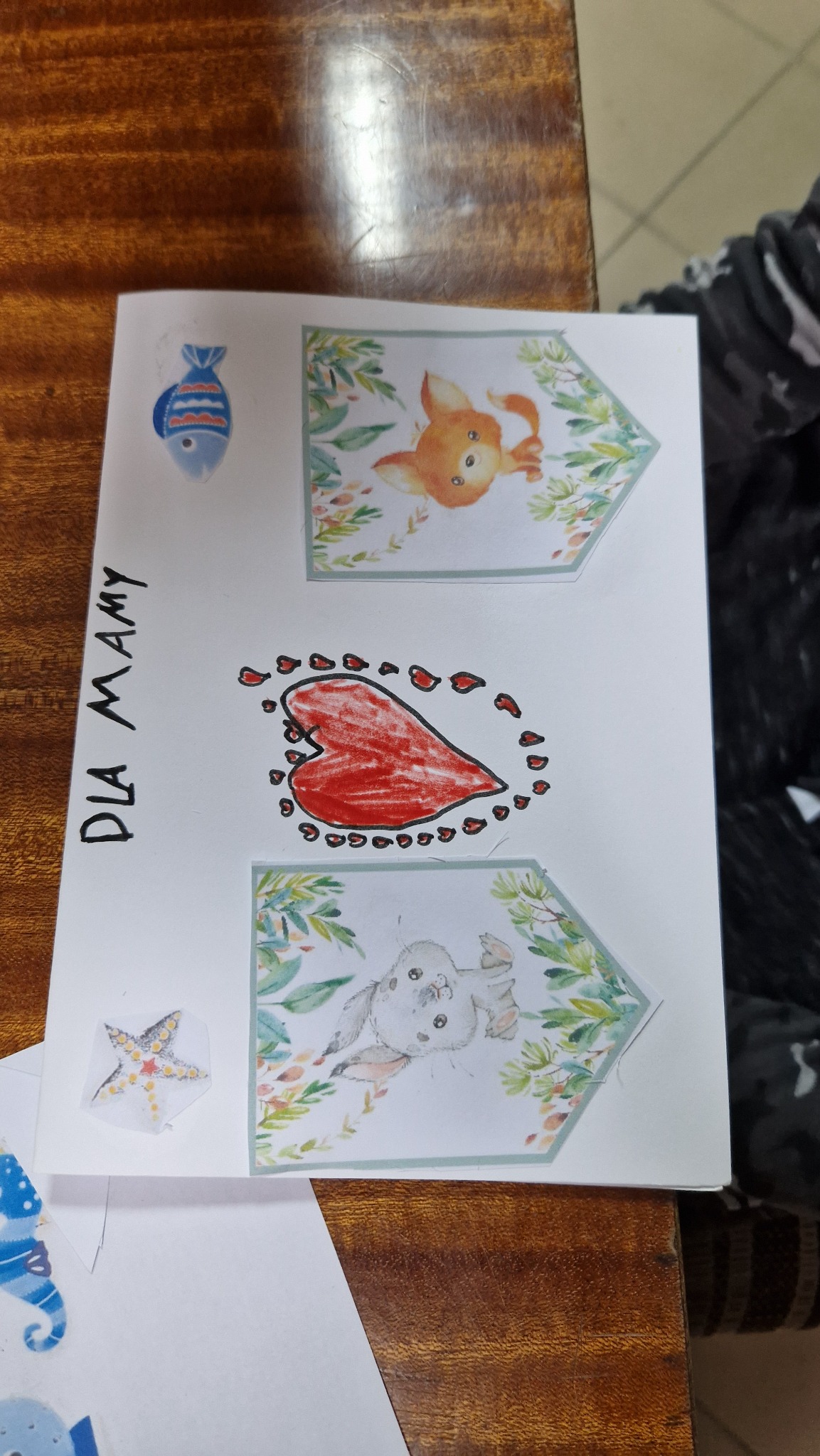 Ręcznie robiona kartka z napisem 'Dla Mamy', ozdobiona naklejkami ryby, gwiazdy oraz rysunkami zwierząt w ramkach z motywem roślinnym: lisa, serca i królika.